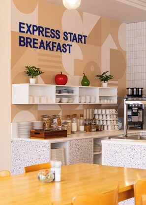 Un espace réservé au petit-déjeuner, avec une pancarte indiquant « PETIT-DÉJEUNER EXPRESS », des étagères remplies d’assiettes, de bols et de bocaux, une machine à café et une table en bois avec des chaises.