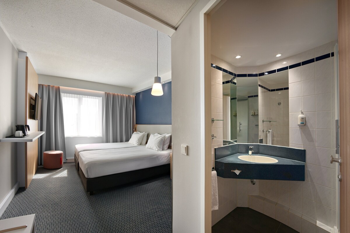 A hotel room with a bed, bedside table, and a bathroom with a sink and mirror.