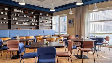 Un comedor moderno con mesas de madera y sillas azules, un banco acolchado azul y estantes con vajilla y copas.
