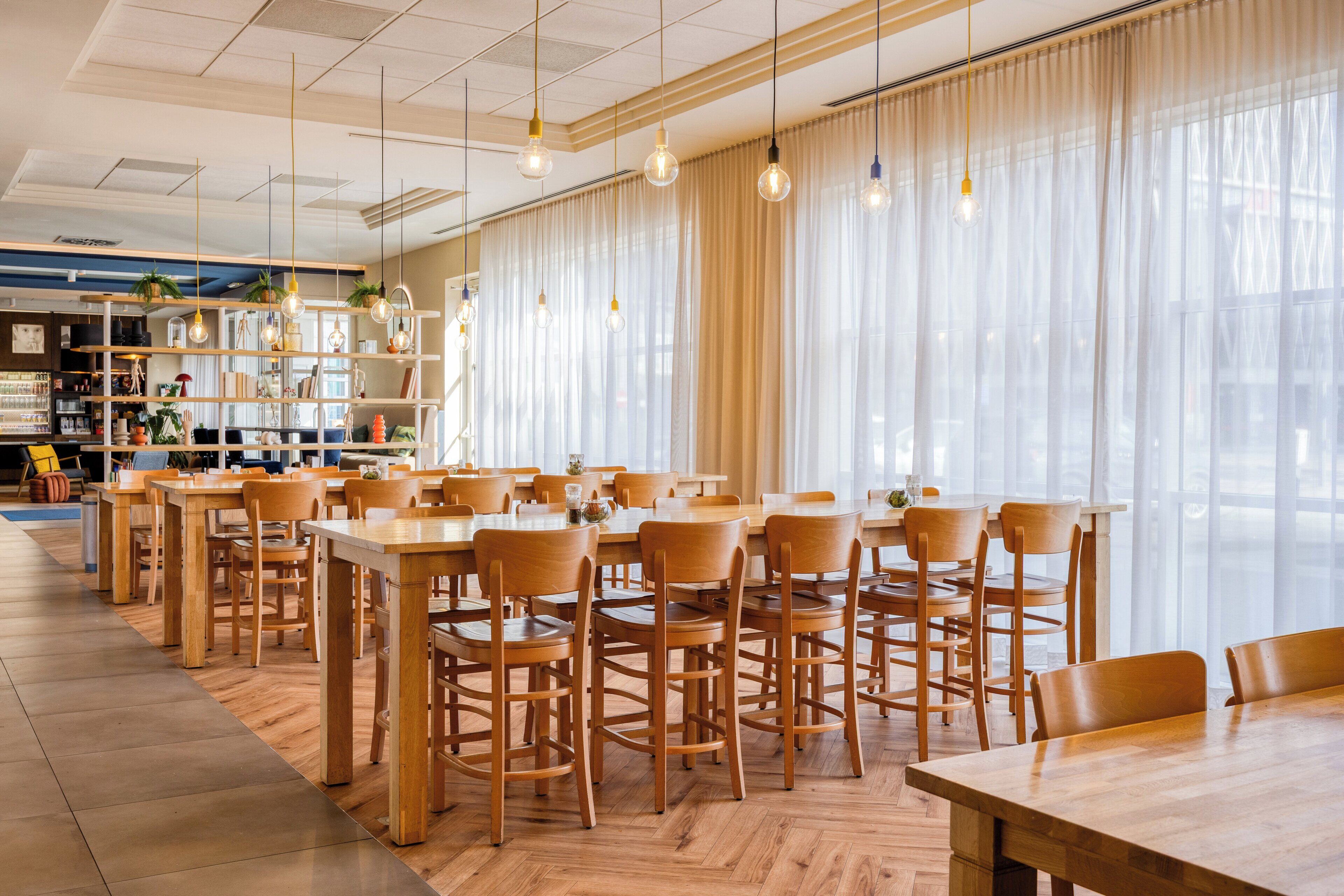 Un comedor amplio con mesas y sillas de madera, ventanas grandes con cortinas translúcidas y lámparas colgantes.