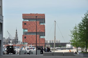 Un bâtiment moderne à la façade distinctive, aux couleurs rouge et marron, situé en bord de mer, avec des bateaux à proximité, et une tour historique en arrière-plan.