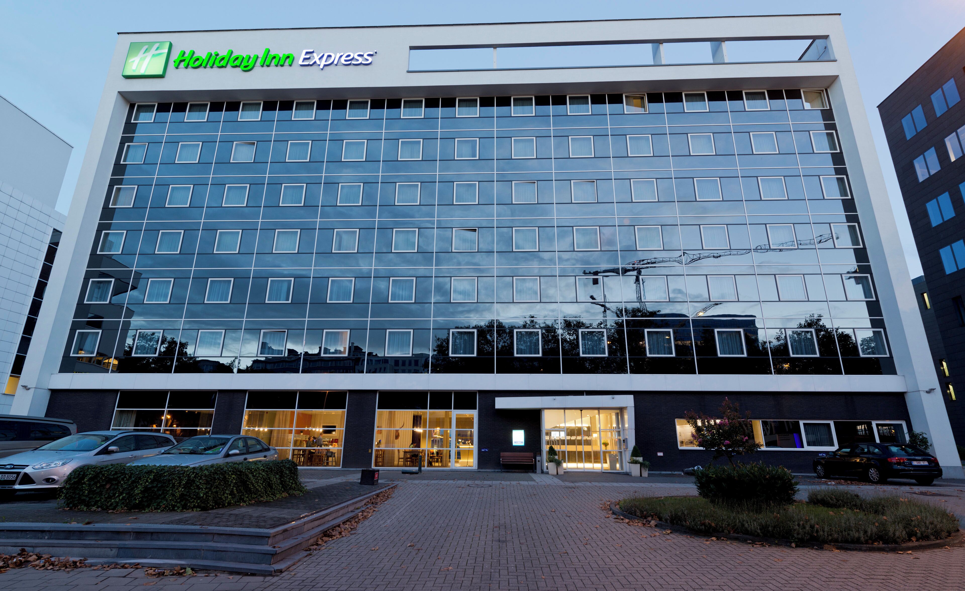 Een Holiday Inn Express hotel met een moderne glazen gevel, een entree van baksteen en een parkeerplaats met auto's.