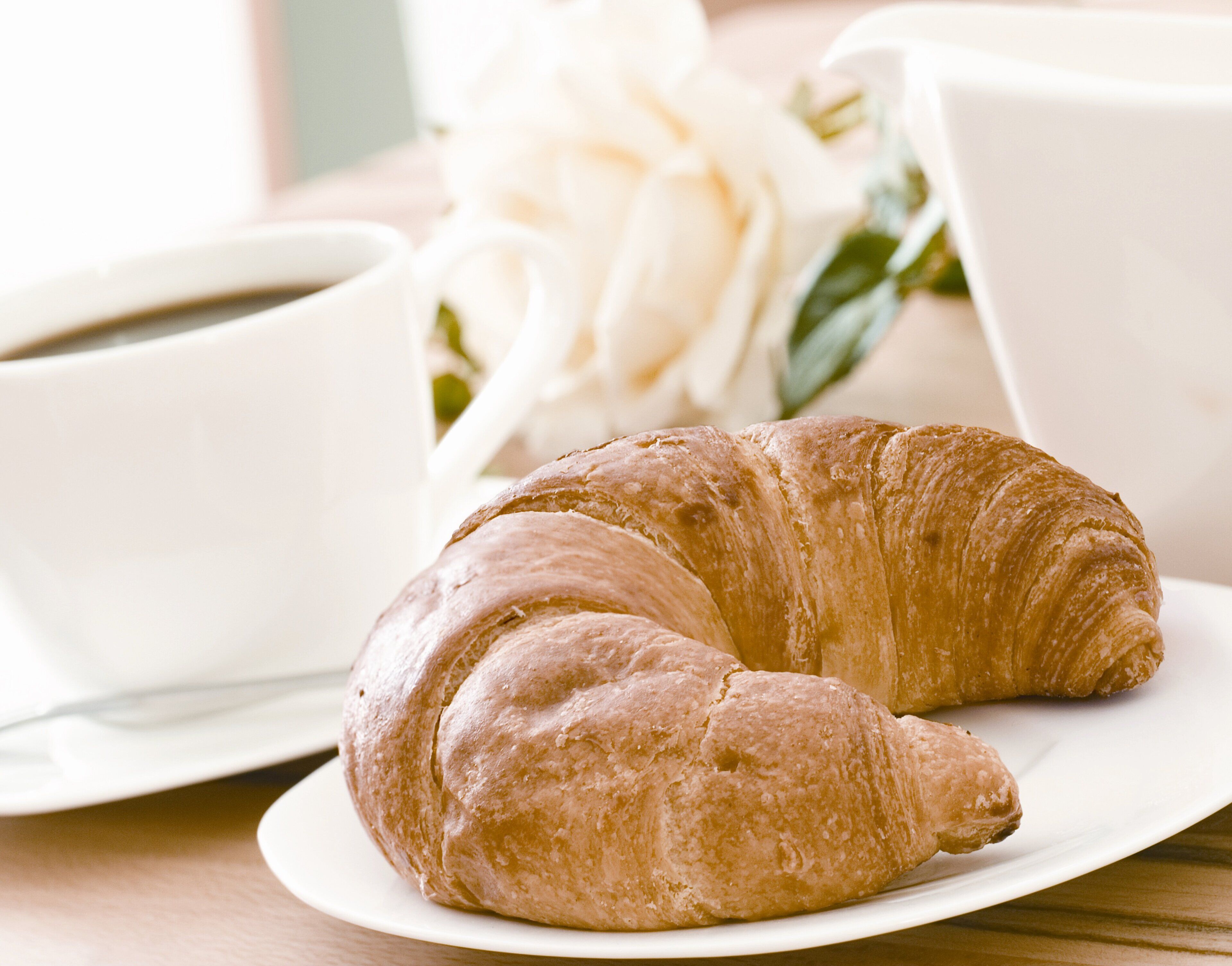 Een croissant op een bord met een kop koffie en een boeket witte bloemen.