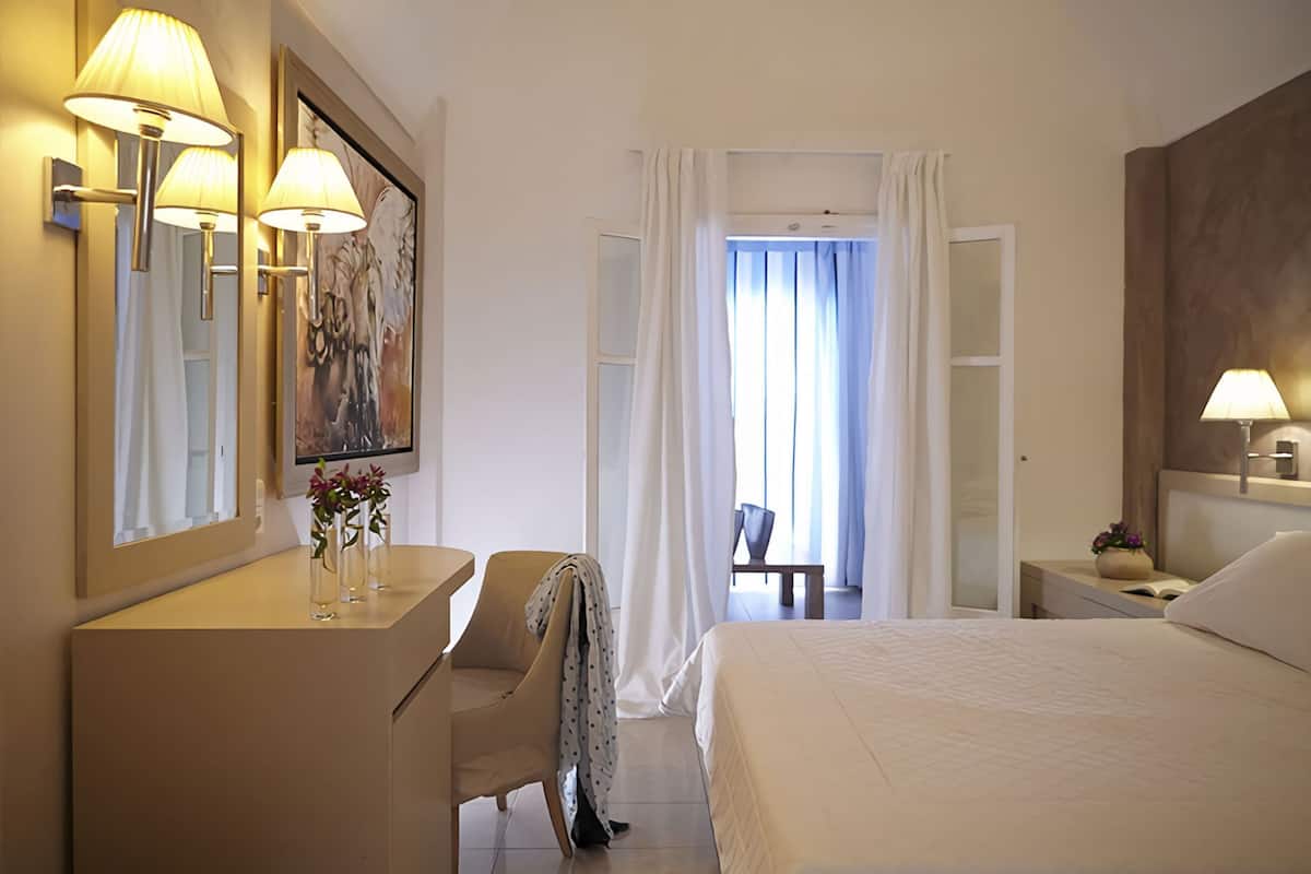 A hotel room with a bed, a desk with lamps, a chair, a painting, and a view of the outdoors through a window.