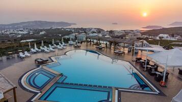 A swimming pool with sun loungers and umbrellas, surrounded by a view of a coastal town during sunset.