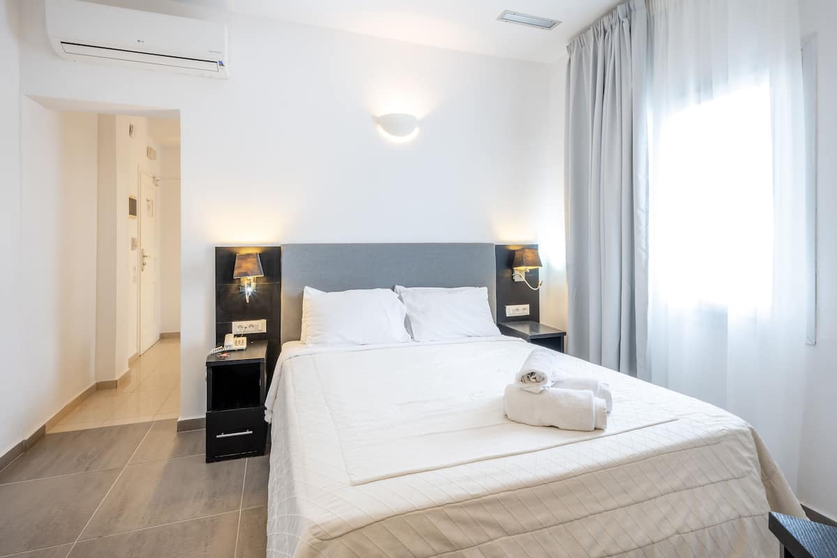 A hotel room with a large bed, two bedside tables with lamps, a wall-mounted light, a door, and a window with curtains.