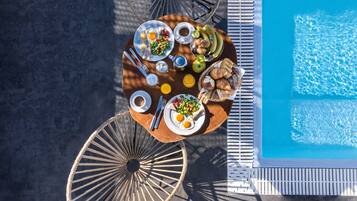 An outdoor dining area with a pool, featuring a round table with plates of food, utensils, and a wicker chair.