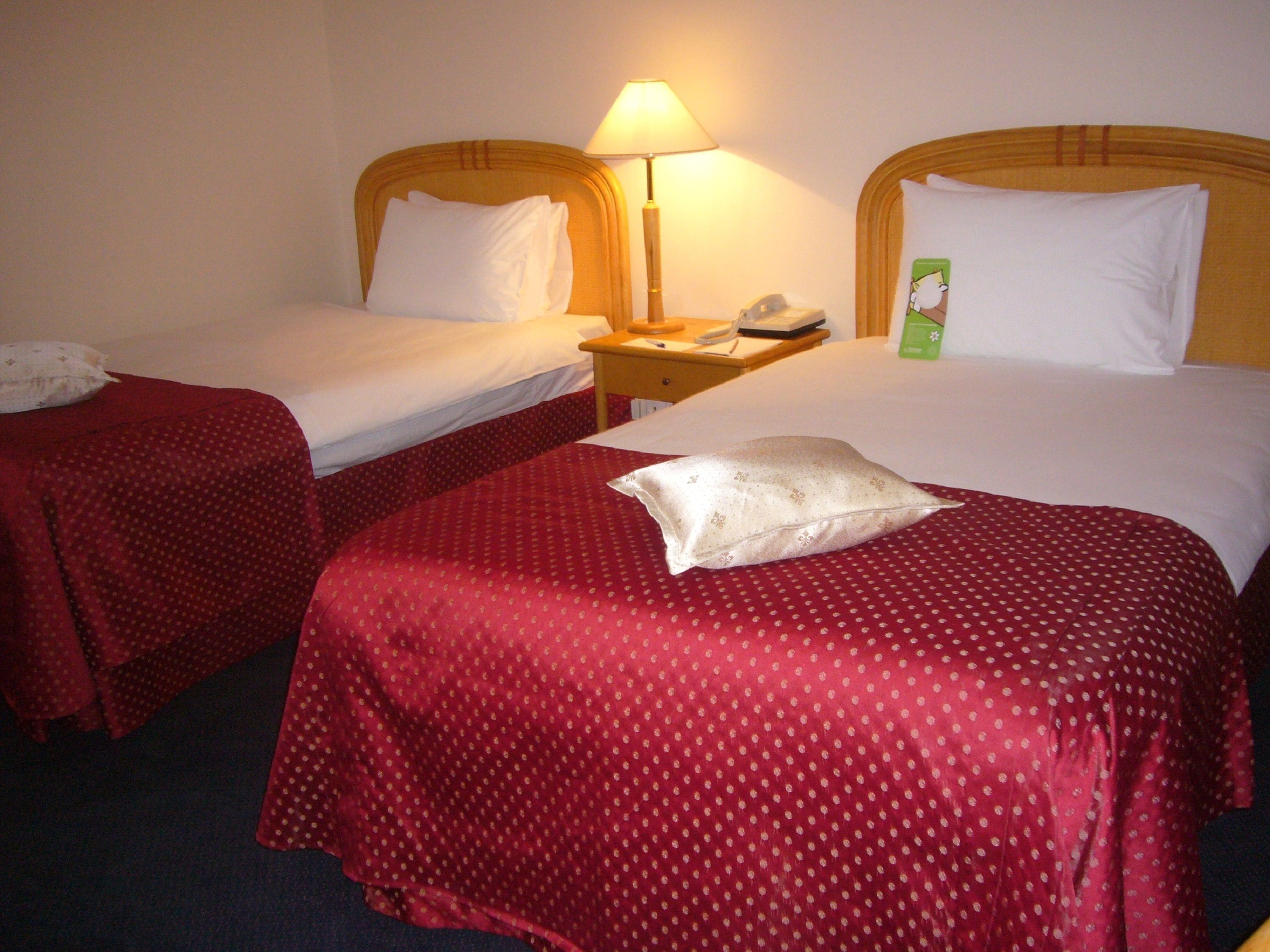 Een hotelkamer met twee eenpersoonsbedden, elk met een rode sprei en witte kussens. Er staat een nachtkastje met een lamp en een telefoon.