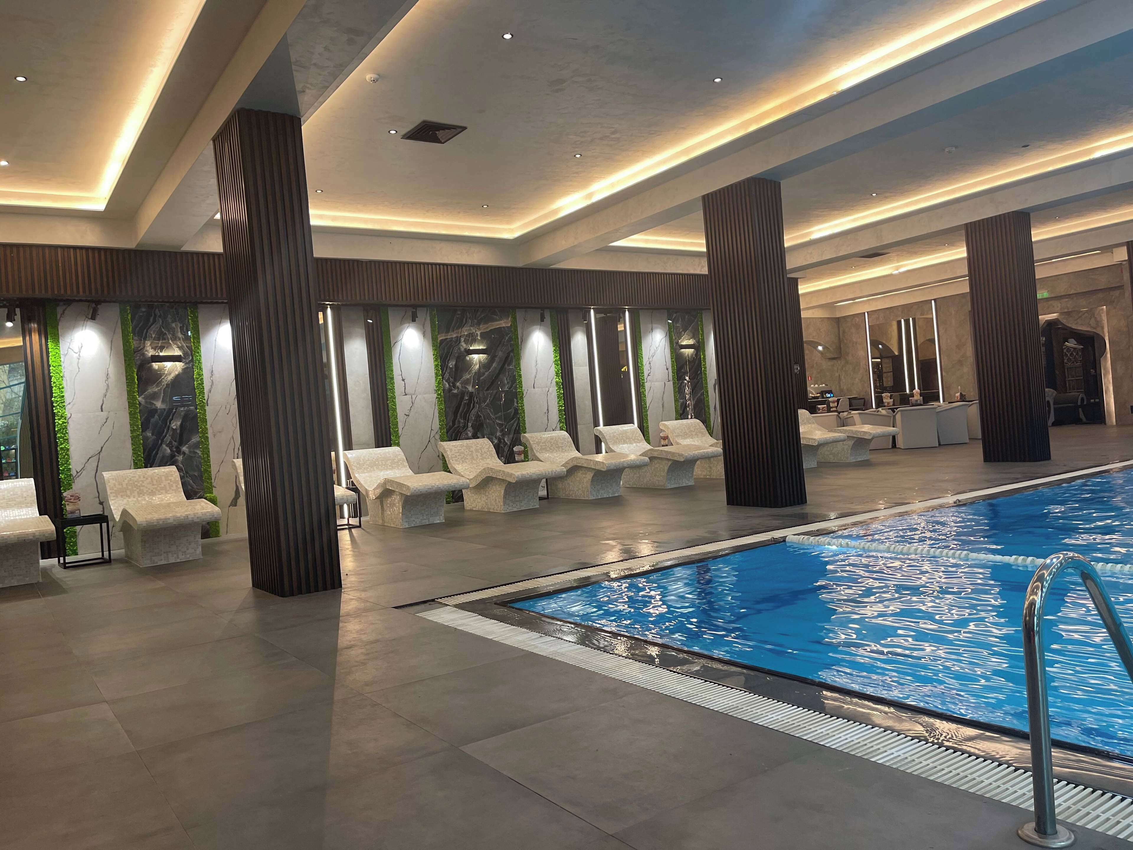 A modern indoor swimming pool area with lounge chairs, a pool, and a reception desk.
