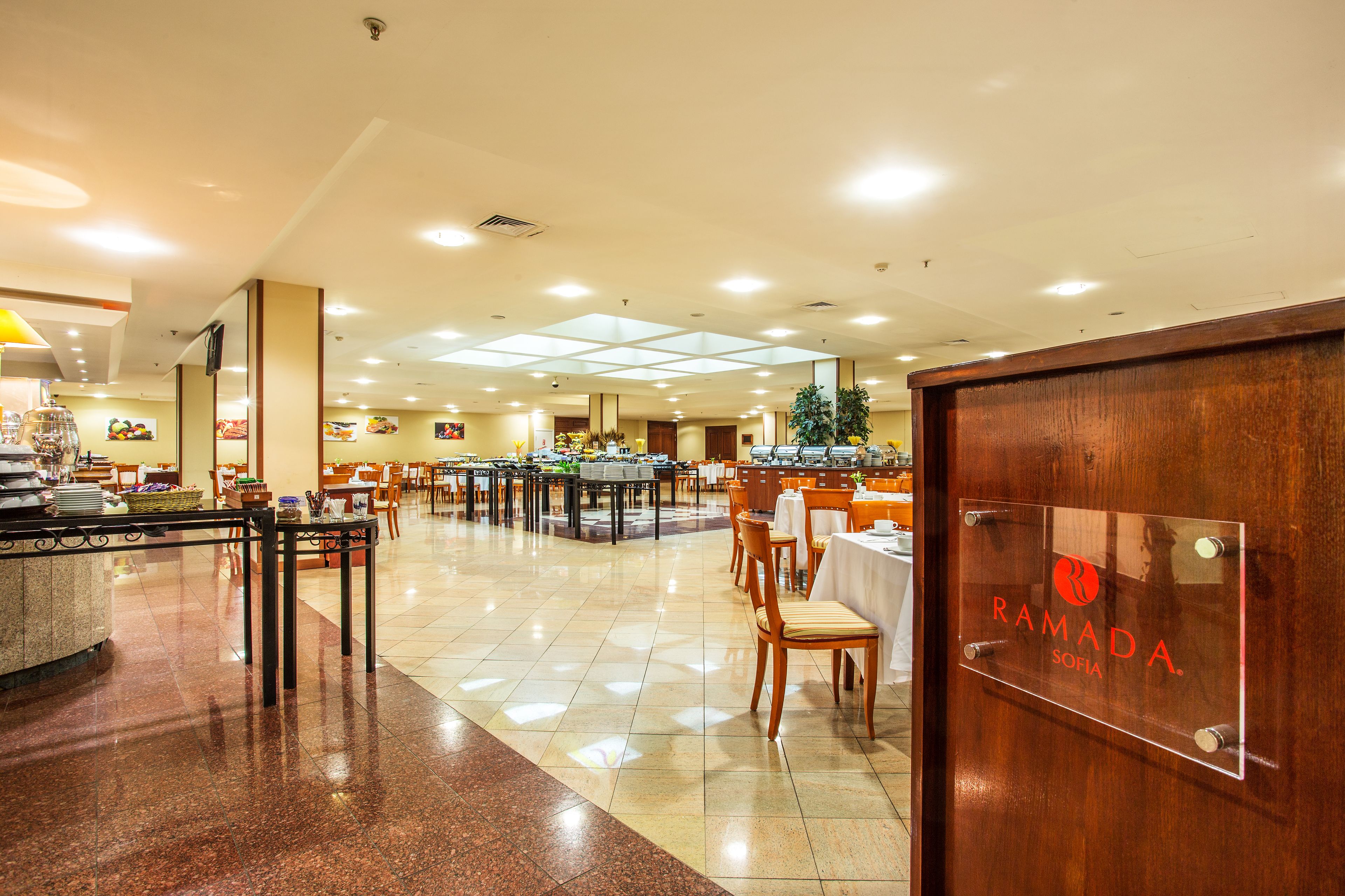 A spacious hotel lobby with multiple dining areas, a reception desk, and a sign indicating the hotel name.