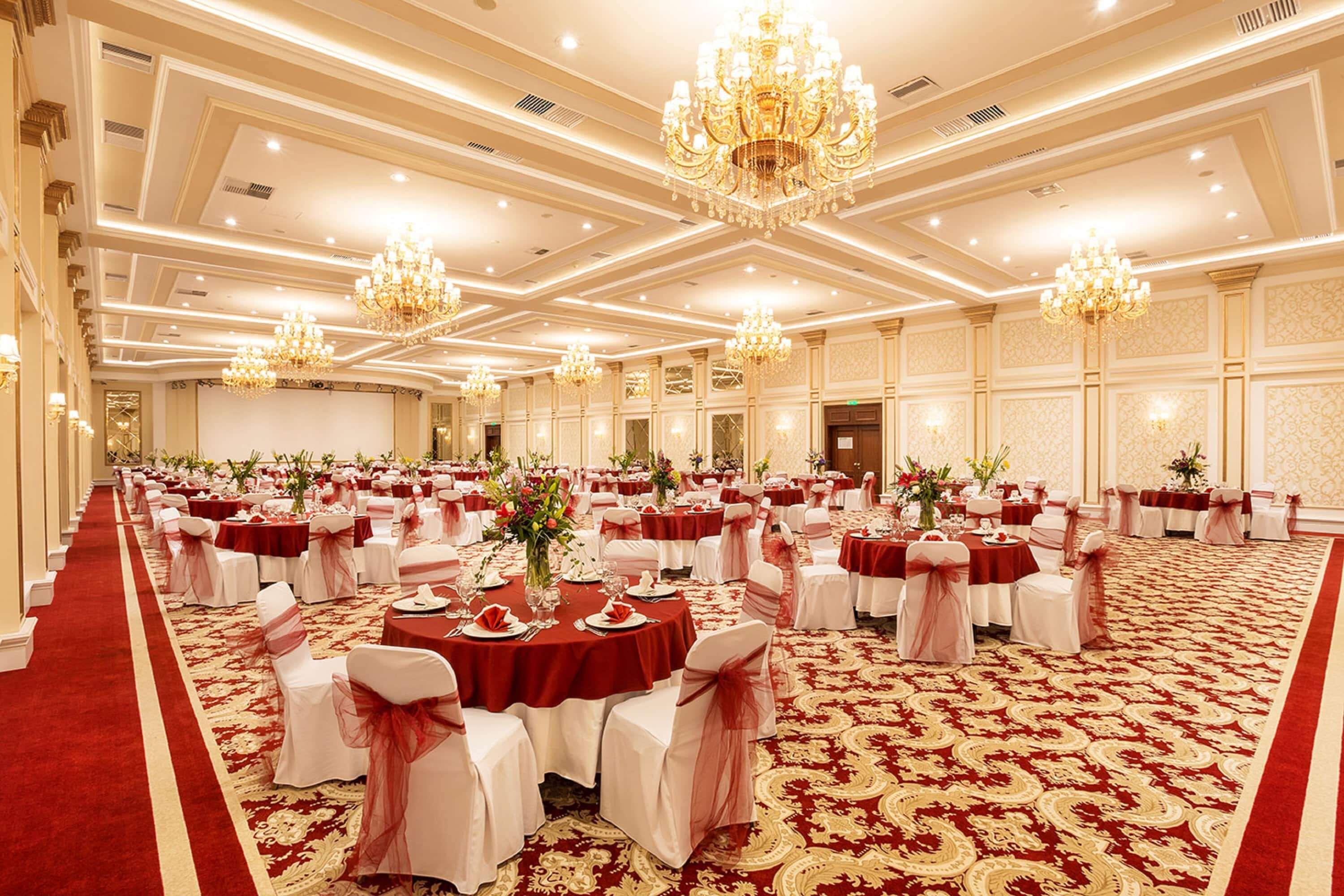 A banquet hall with round tables, red carpets, and chandeliers.