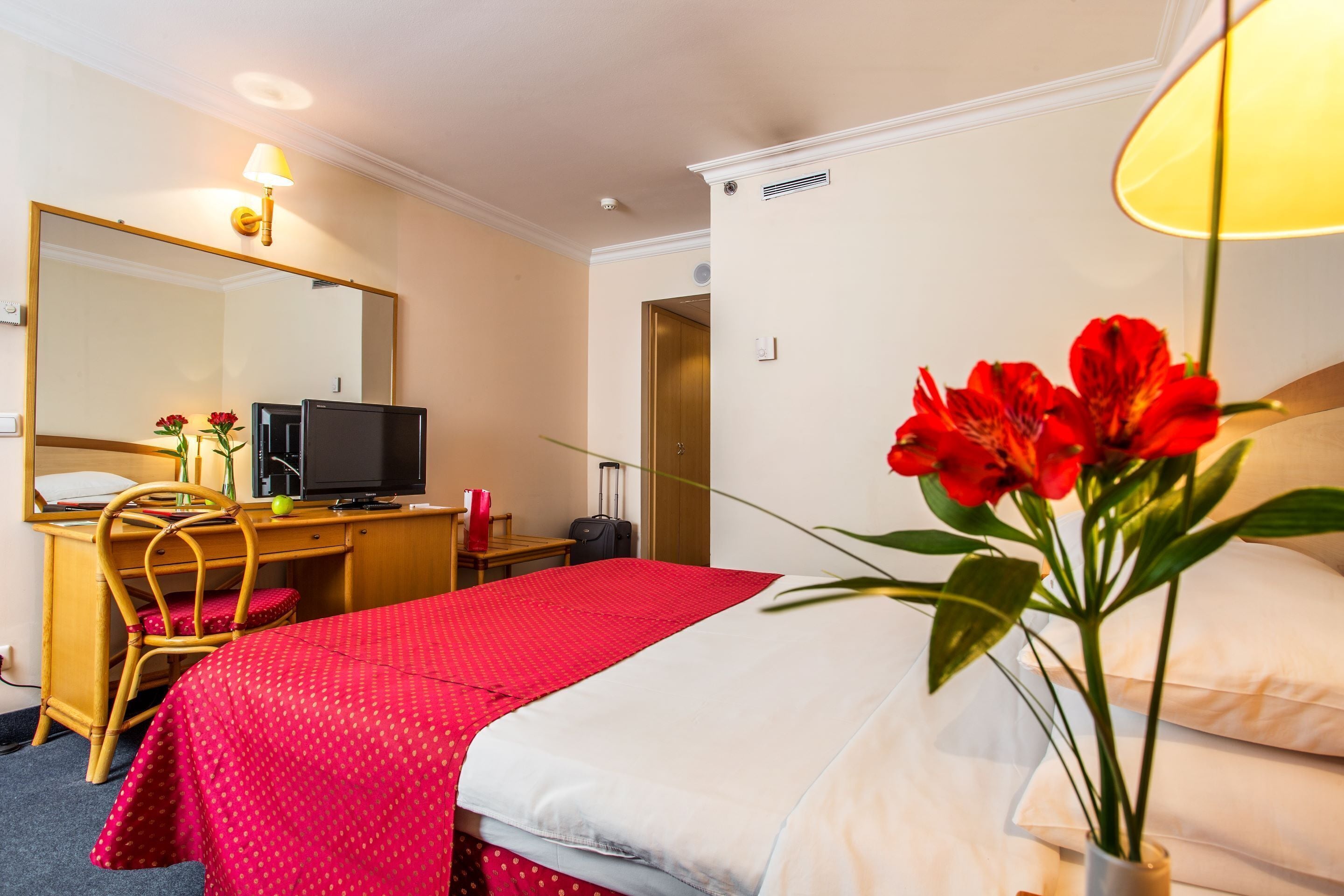 A hotel room with a bed, a desk with a computer, a chair, and a large mirror.