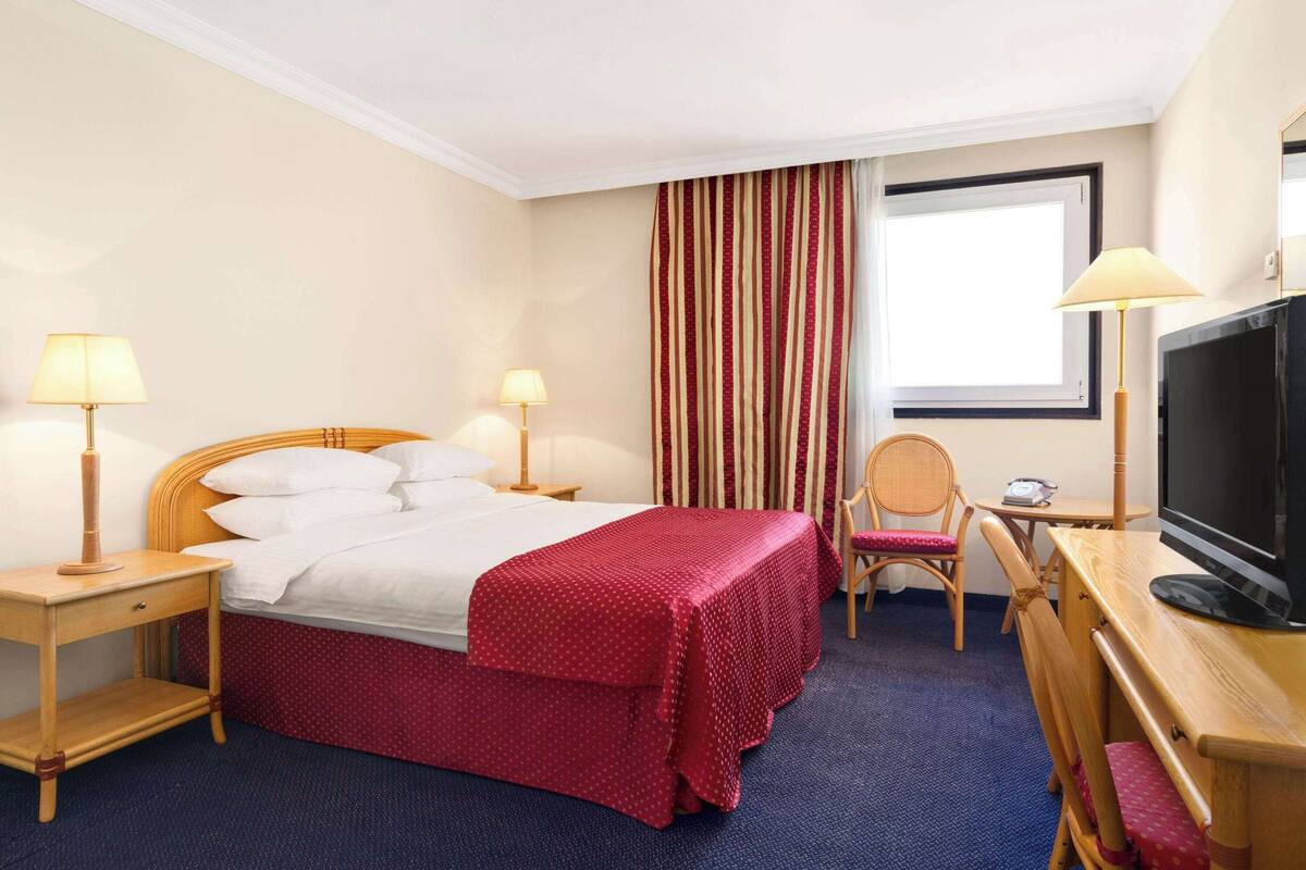 Une chambre d’hôtel avec un lit, deux tables de chevet avec des lampes, un téléviseur, une chaise et une fenêtre avec des rideaux.