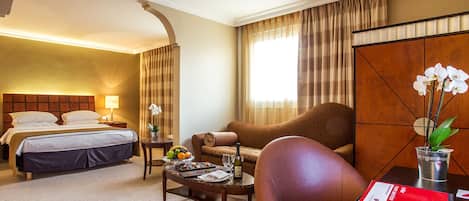 A hotel room with a bed, a sofa, a coffee table, and a dining table. There is a window with curtains, a lamp, and a plant.