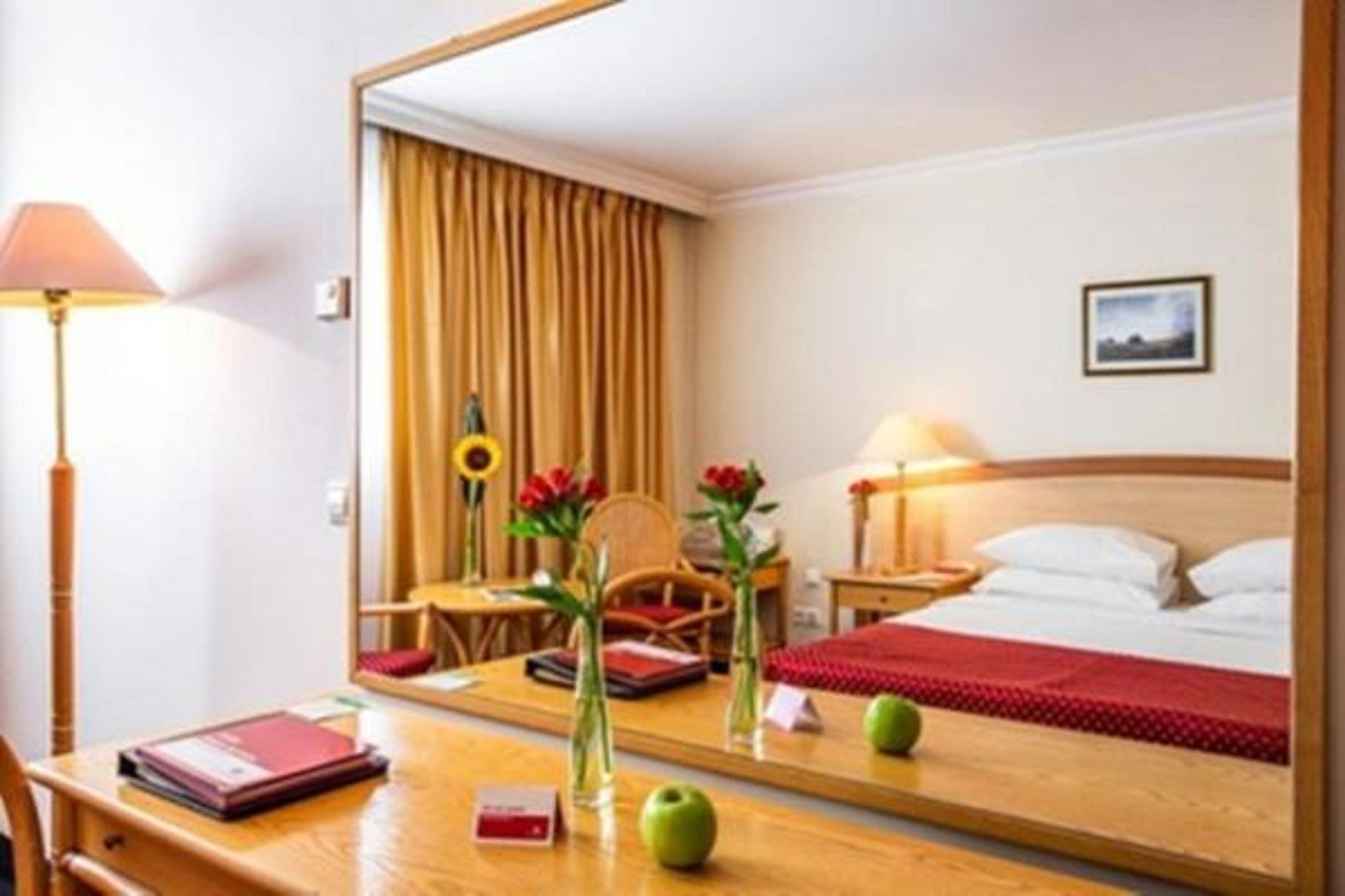 A hotel room with a bed, a desk with a lamp, a chair, a mirror, and a window with curtains.