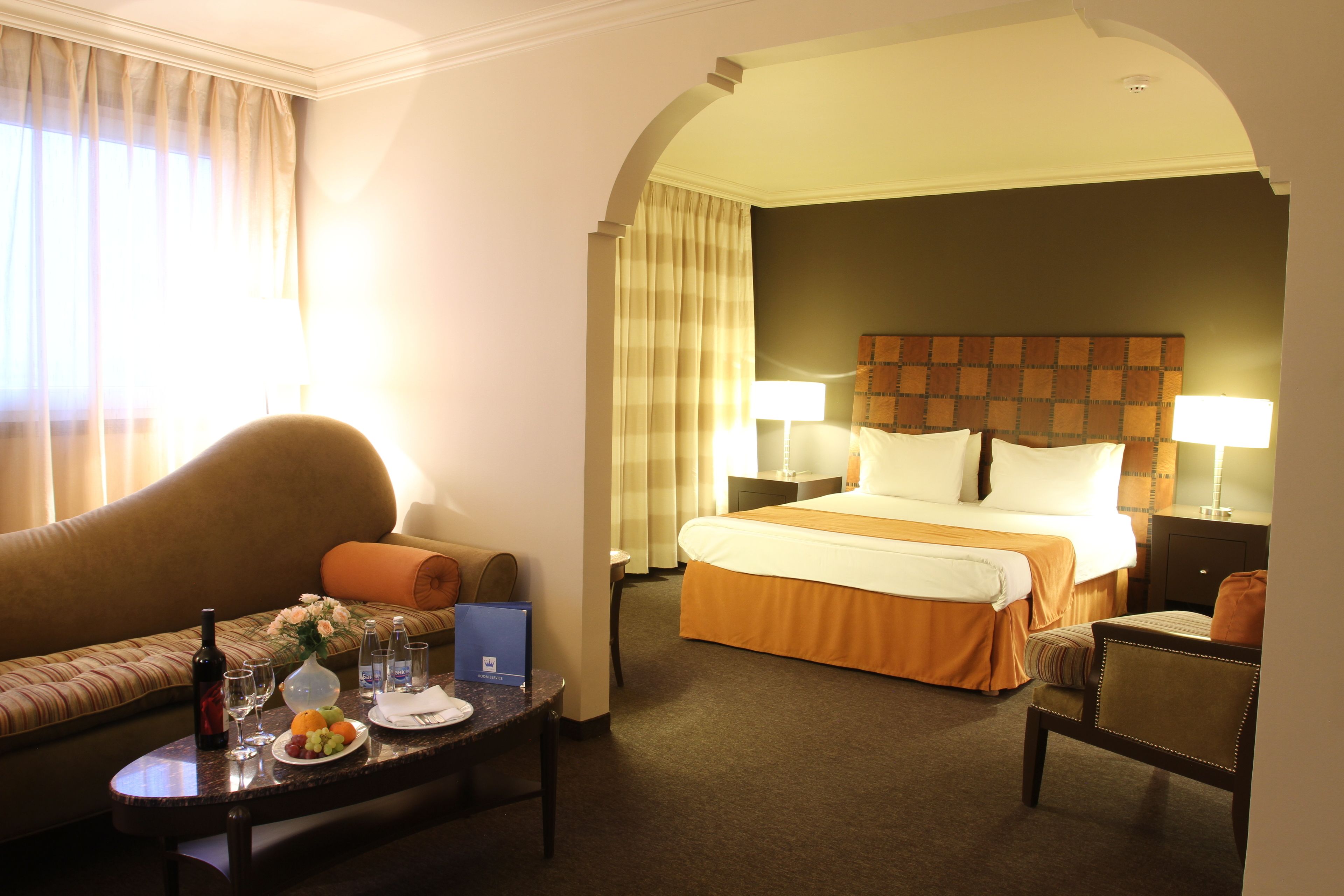 A hotel room with a bed, two bedside tables, a sofa, a small table with a fruit plate, a bottle of wine, and a vase of flowers.
