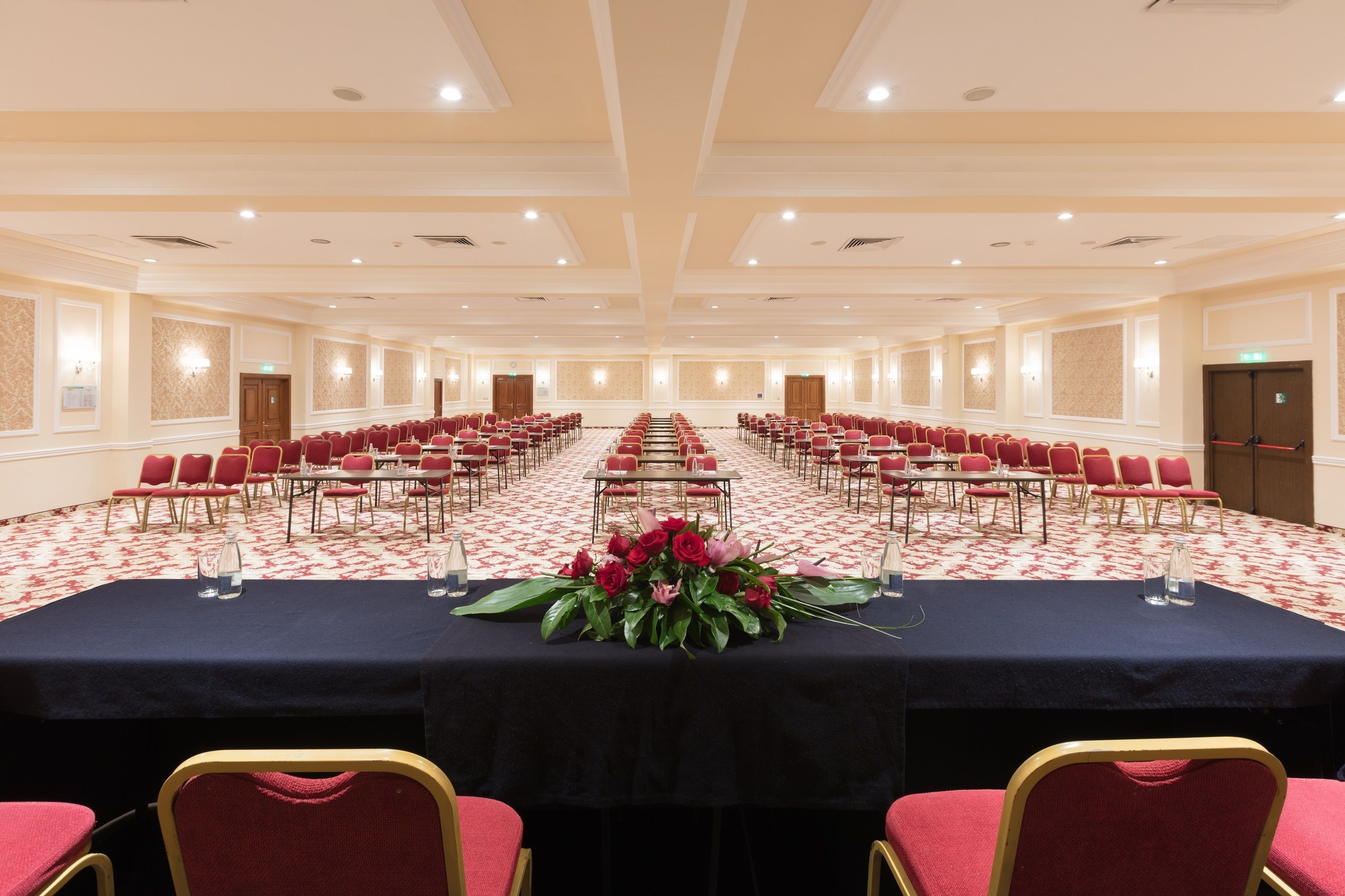 Un salón de conferencias con filas de sillas rojas, una mesa larga con mantel negro y un centro de mesa floral.