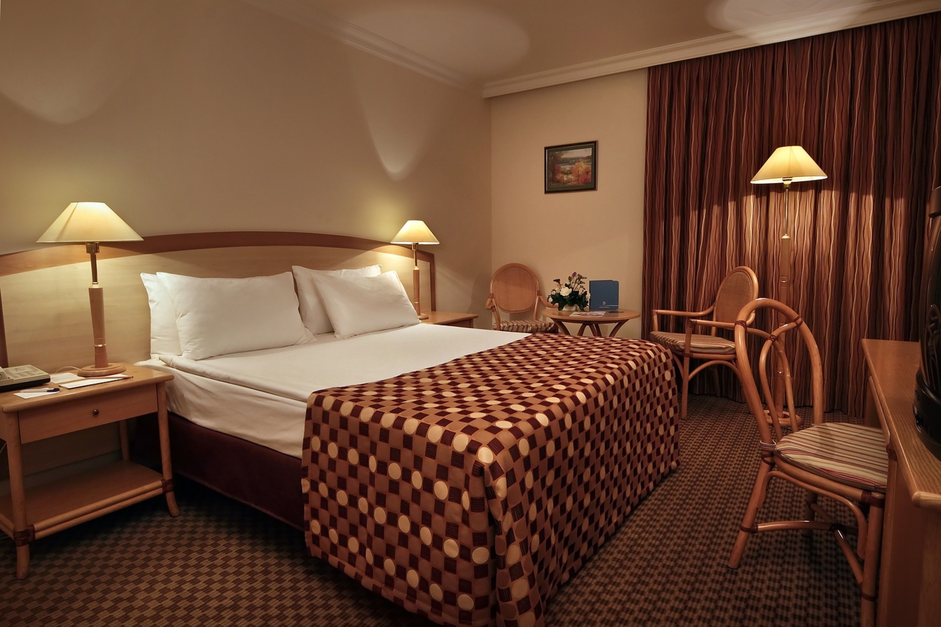 A hotel room with a large bed, two bedside tables with lamps, a desk with a chair, and a checkered area rug.