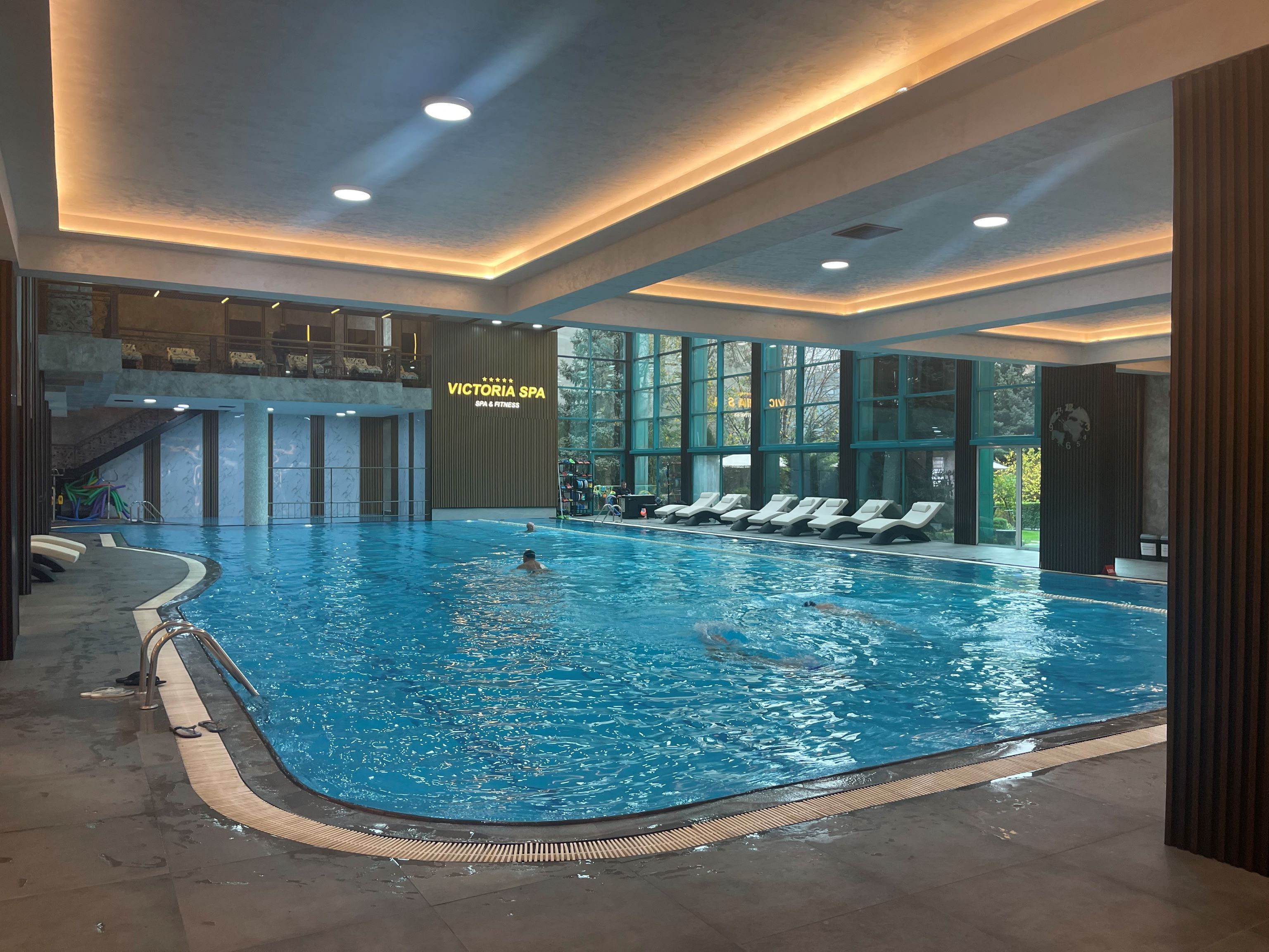 Ein Hallenbad mit klarem Wasser, eingelassenen Liegestühlen und einem Schild mit der Aufschrift "Victoria SPA & FITNESS".