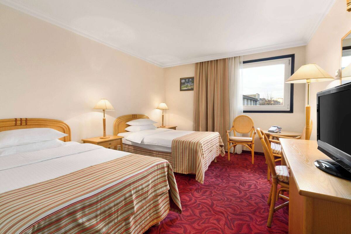 Une chambre d’hôtel avec deux lits, un bureau, une chaise, une télévision et une fenêtre avec des rideaux.