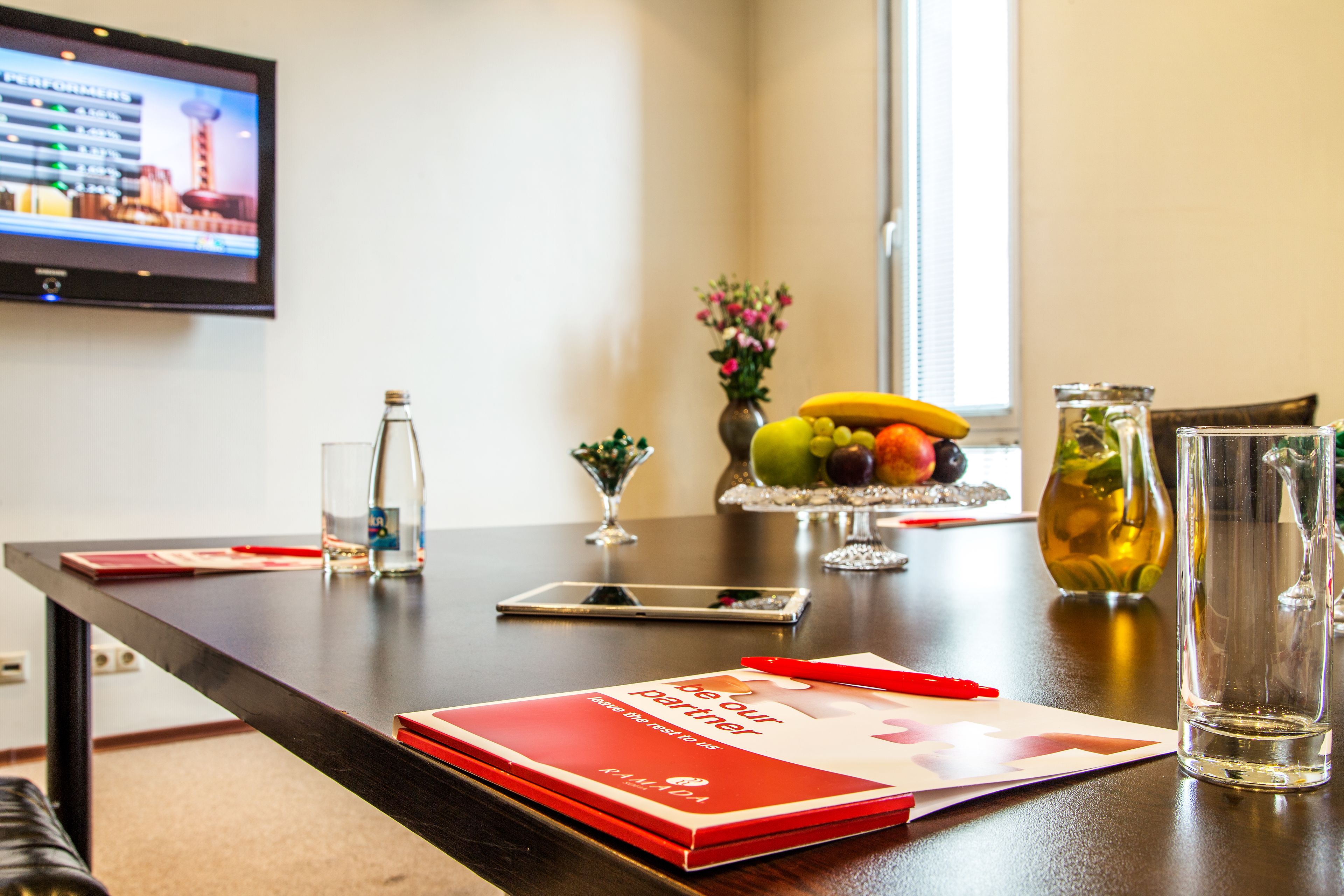 A conference table with a red folder, a smartphone, a glass pitcher, and a fruit bowl.