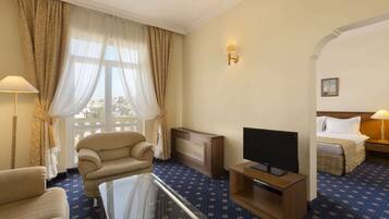 Habitación de hotel con sofá, televisor sobre un mueble bajo, cama con cabecera y ventana con cortinas.