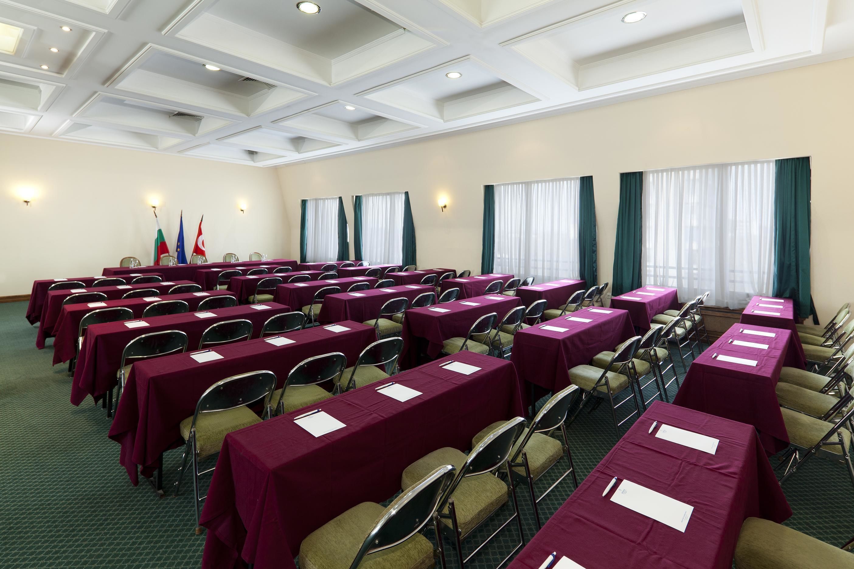 A conference room with rows of tables and chairs, flags, and curtains.