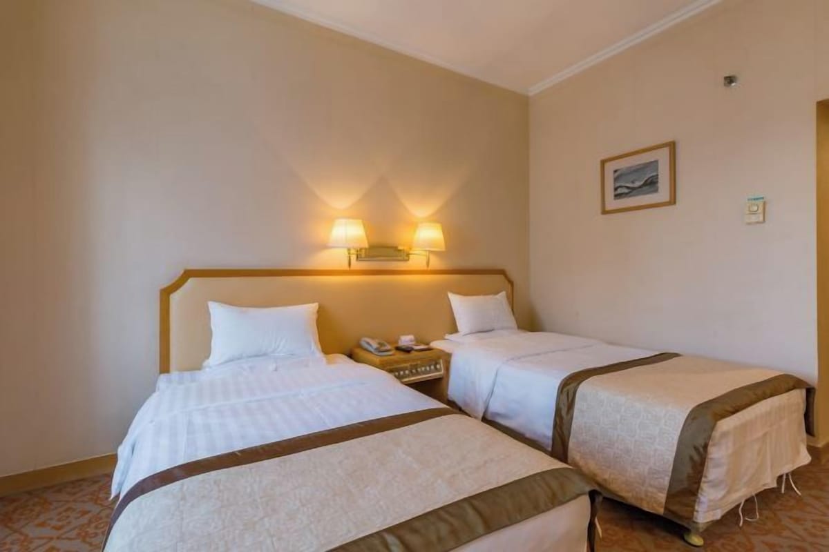 A hotel room with two beds, a nightstand with a phone, and wall-mounted lamps.