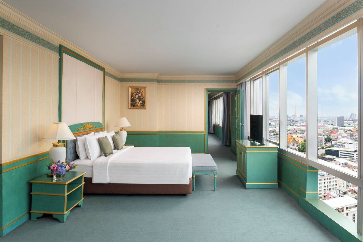 Ein Hotelzimmer mit Bett, Nachttisch, Fernseher und einem großen Fenster mit Blick auf die Stadt.