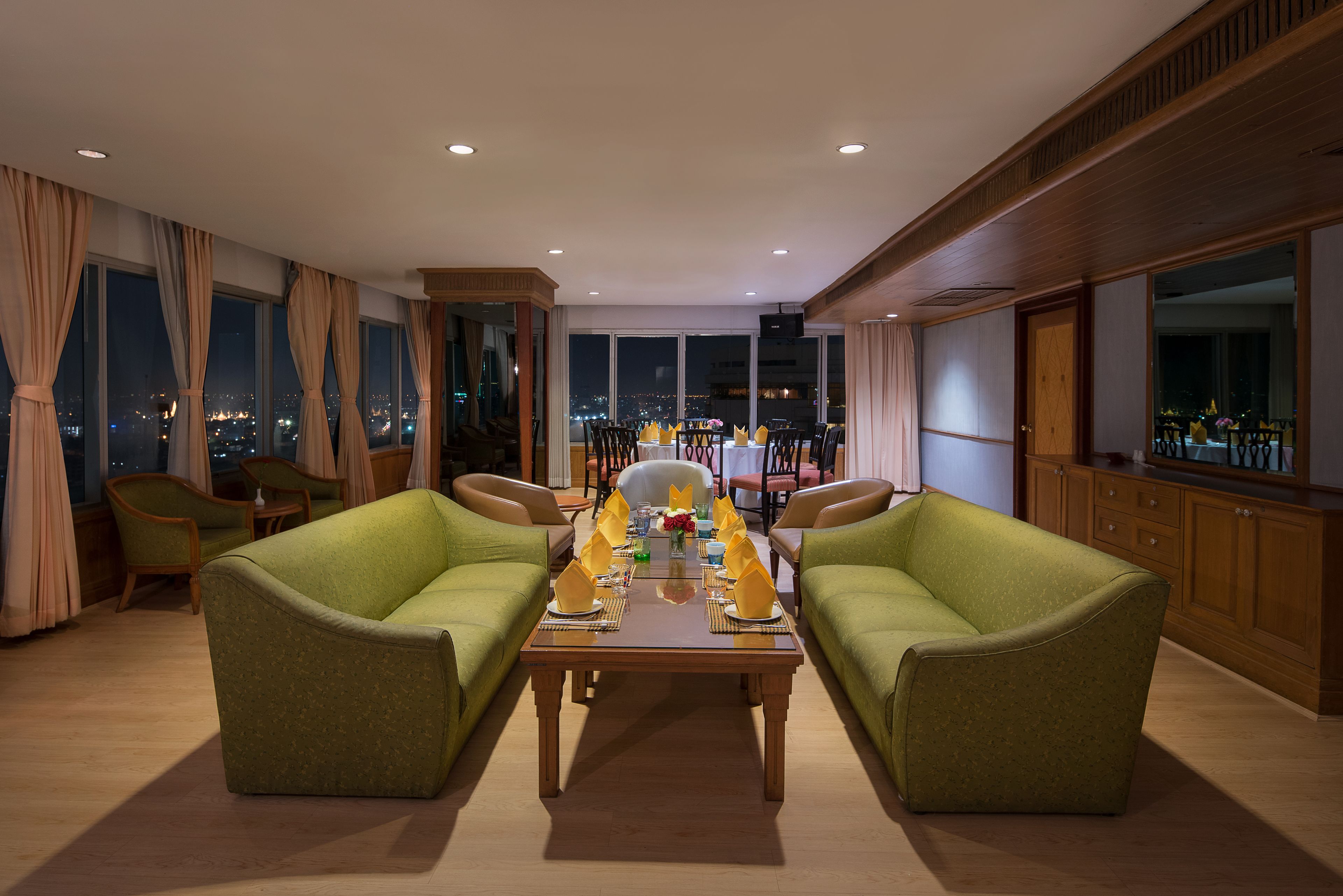 A spacious living room with a large dining table set for six, flanked by two green sofas, large windows with city views, and wooden paneling.