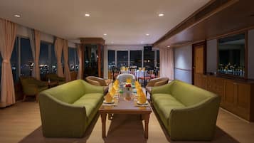 A spacious living room with a large dining table set for six, flanked by two green sofas, large windows with city views, and wooden paneling.