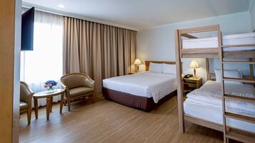 Habitación de hotel con una cama grande, una cama nido de madera, un escritorio con una silla, una mesita con flores y un televisor en la pared.