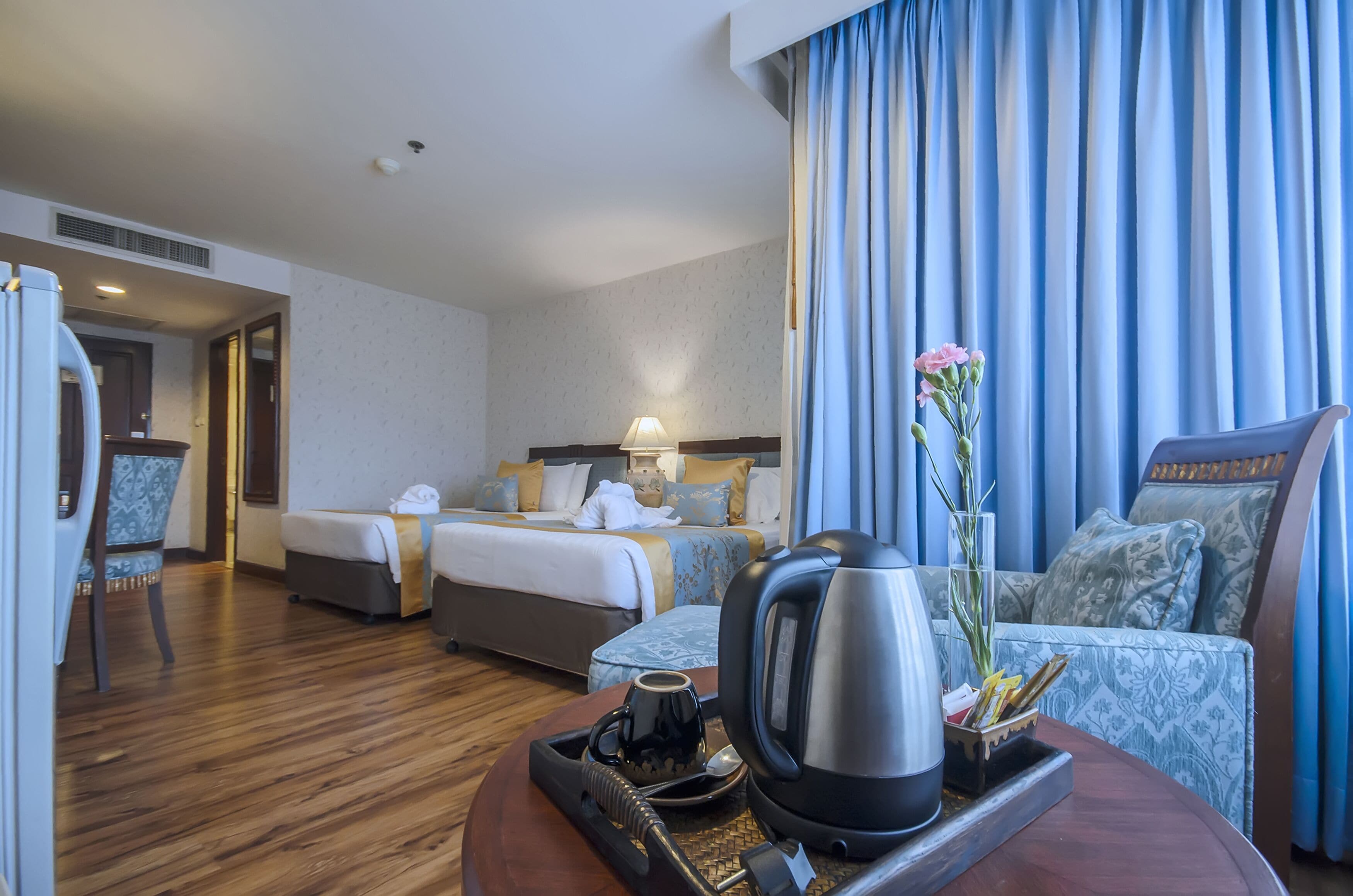 A hotel room with a bed, a sofa, a coffee table with a kettle and a tray, and a vase with flowers.