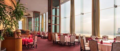 Un comedor de hotel con mesas dispuestas para el servicio, ventanas amplias y suelo alfombrado en rojo.