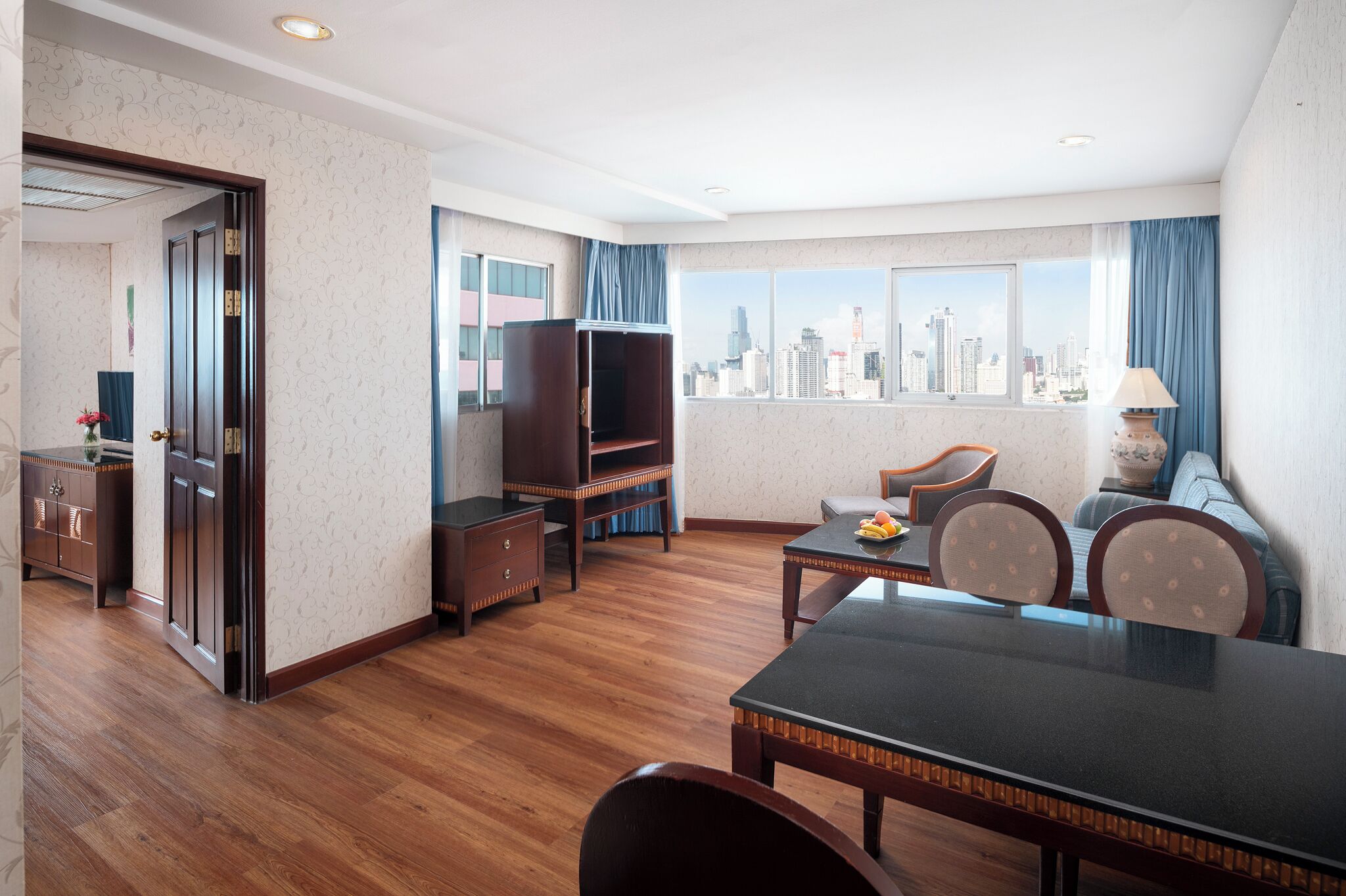 A hotel room with a dining table, chairs, a sofa, a television, and a city view.