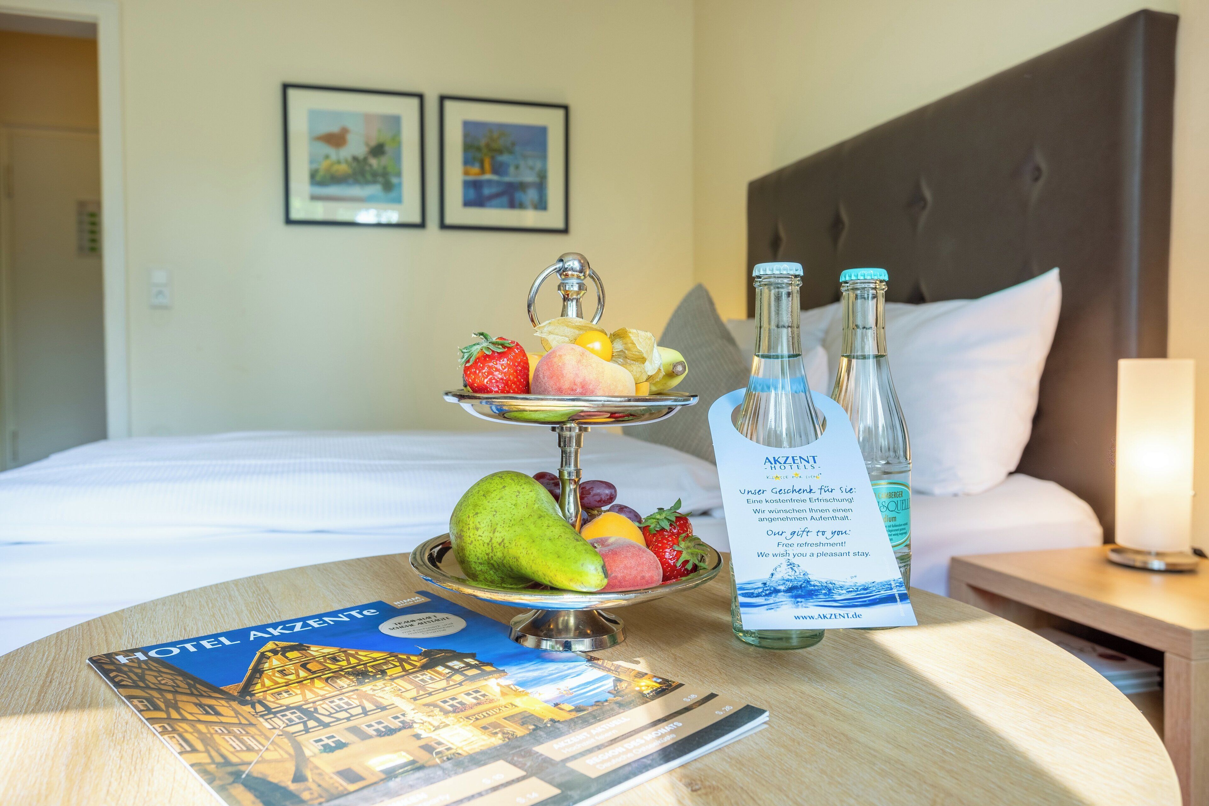 Et hotelværelse med seng, et natbord med frugtfad, en vandflaske og en velkomstfolder.