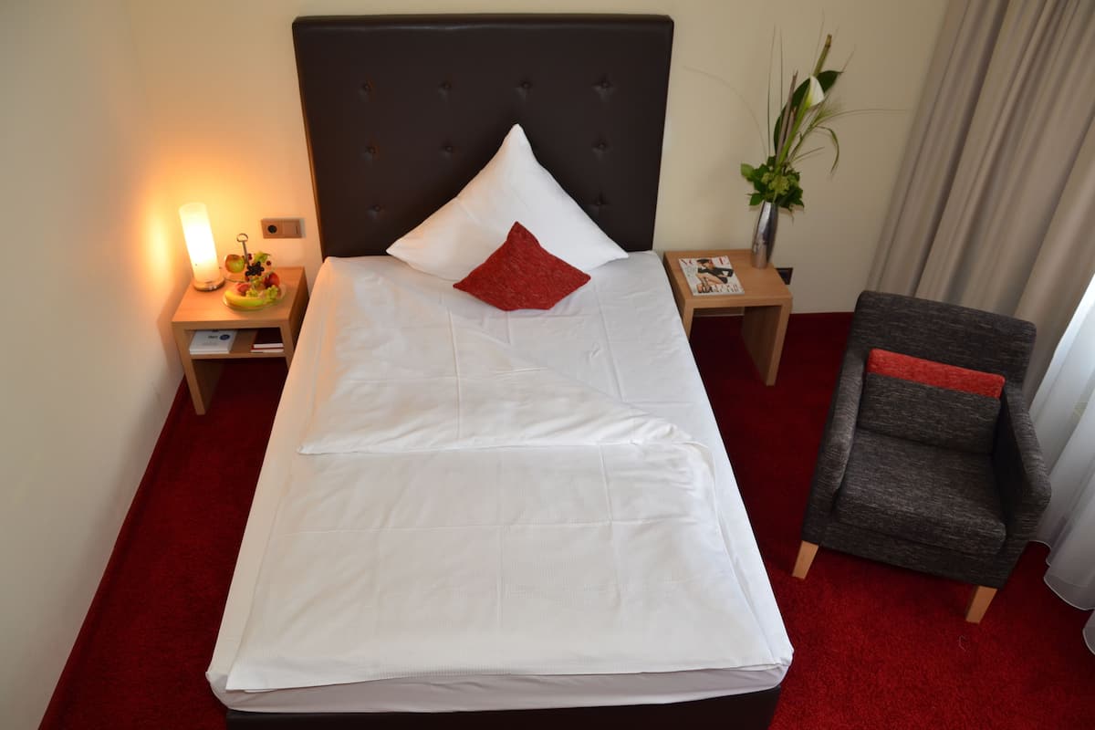 A neatly made bed with white linens and a red pillow, a bedside table with a lamp, a small plant, and a chair with a red cushion.