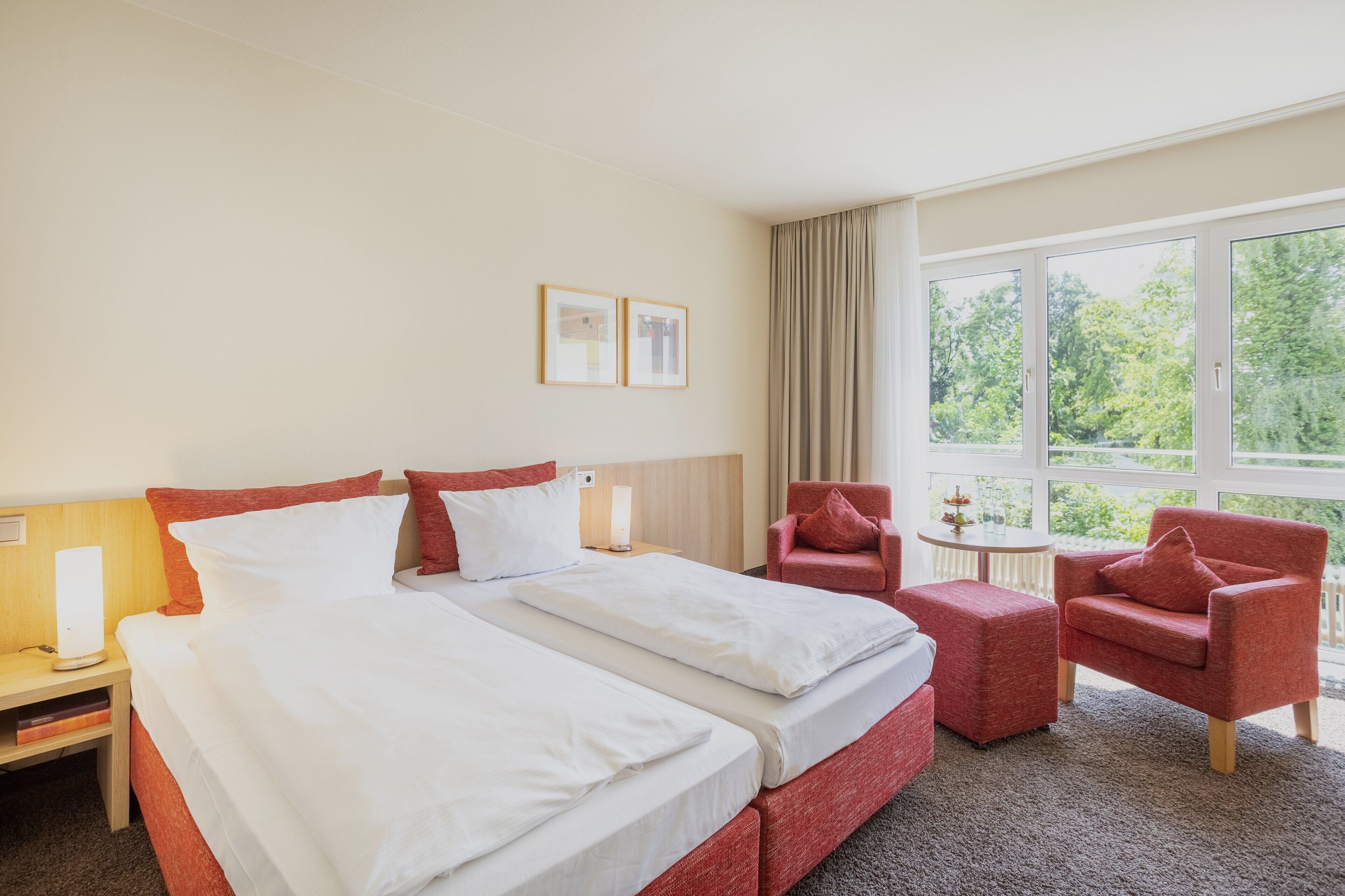 Habitación de hotel con una cama grande, dos sillones rojos, una mesita y una ventana con vistas a árboles.
