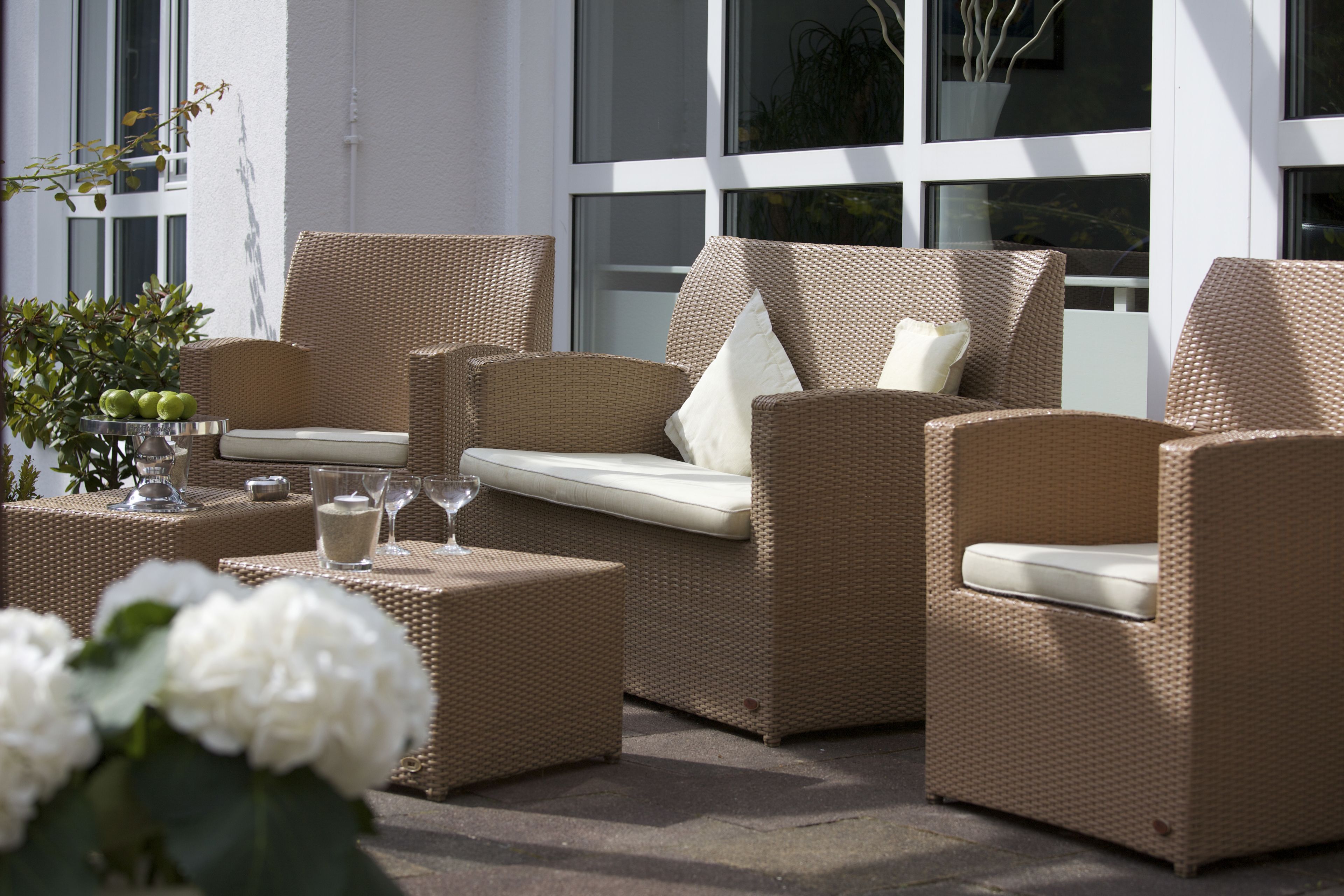Een zithoek buiten van rieten meubels met comfortabele stoelen en een kleine tafel, aangevuld met een potplant en een bloevas.