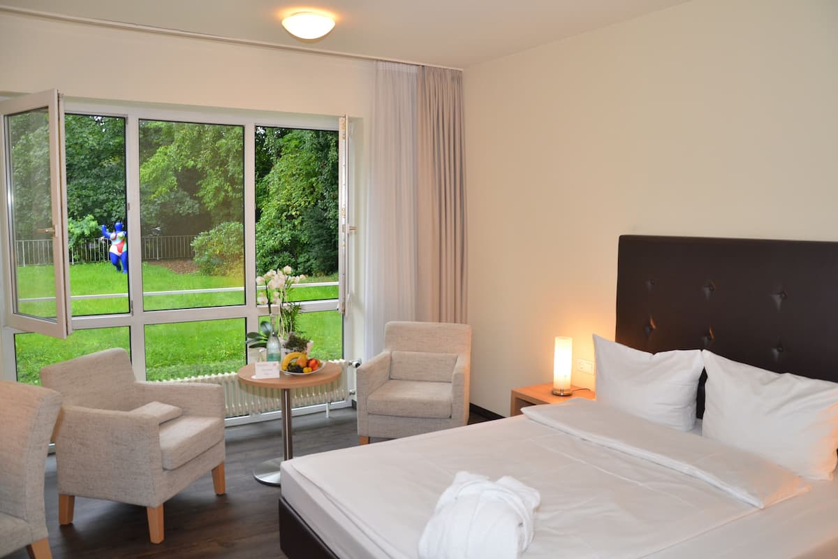 A hotel room with a large bed, two armchairs, a small table with a fruit bowl, and a view of a garden through a large window.