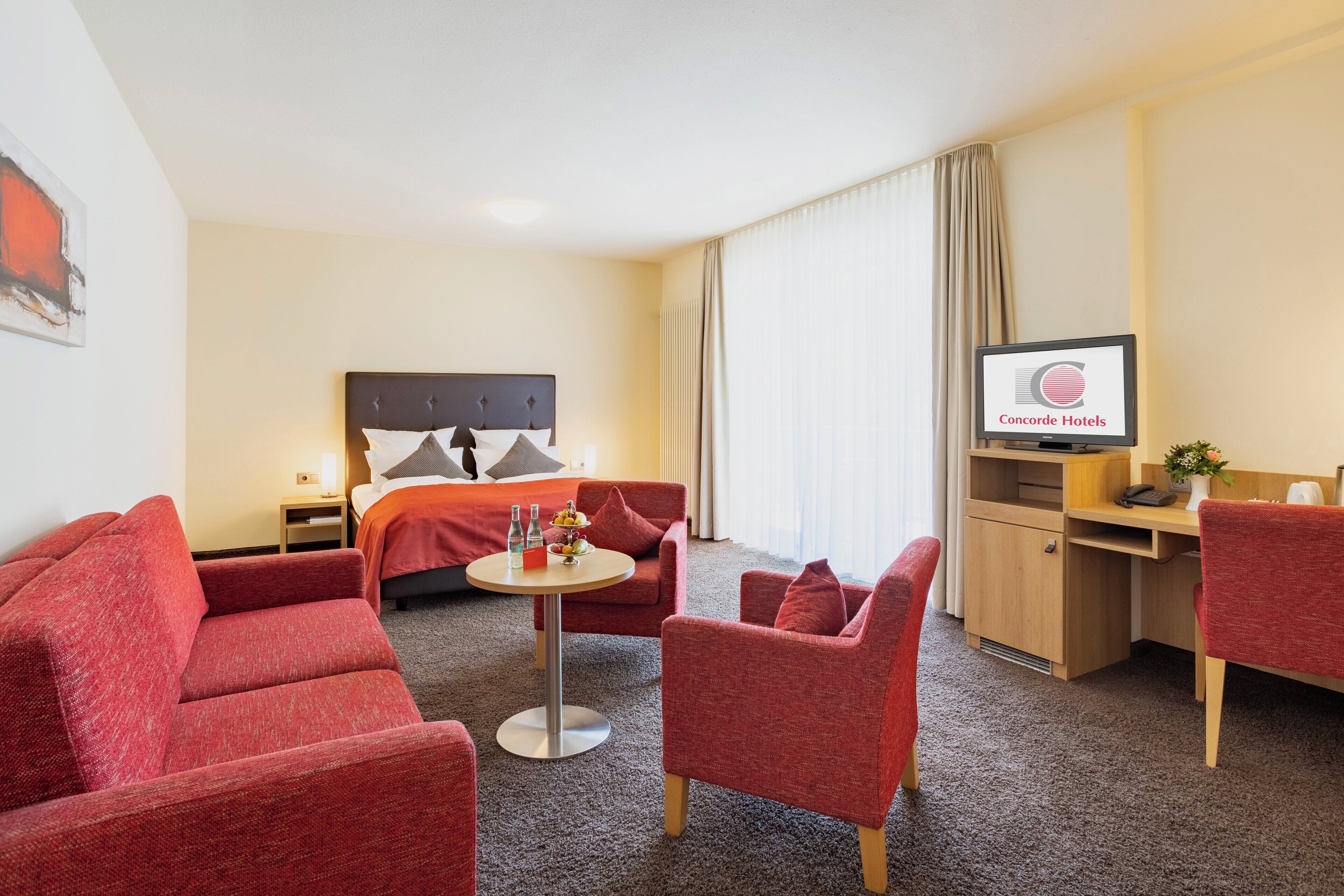 A hotel room with a bed, a desk, a TV, and red upholstered furniture.