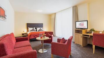 A hotel room with a bed, a desk, a TV, and red upholstered furniture.