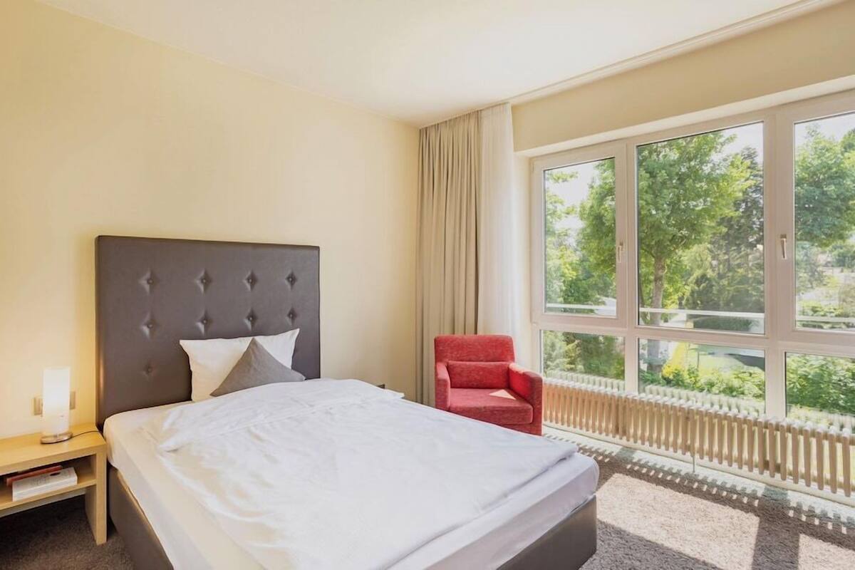 A hotel room with a bed, a nightstand, a red chair, and a large window with a view of greenery.