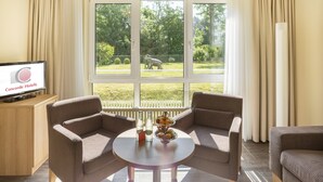Een hotelkamer met twee sofa’s gericht op een groot raam, een bijzettafeltje met een schaal fruit en flessen, en een televisie op een houten kast.