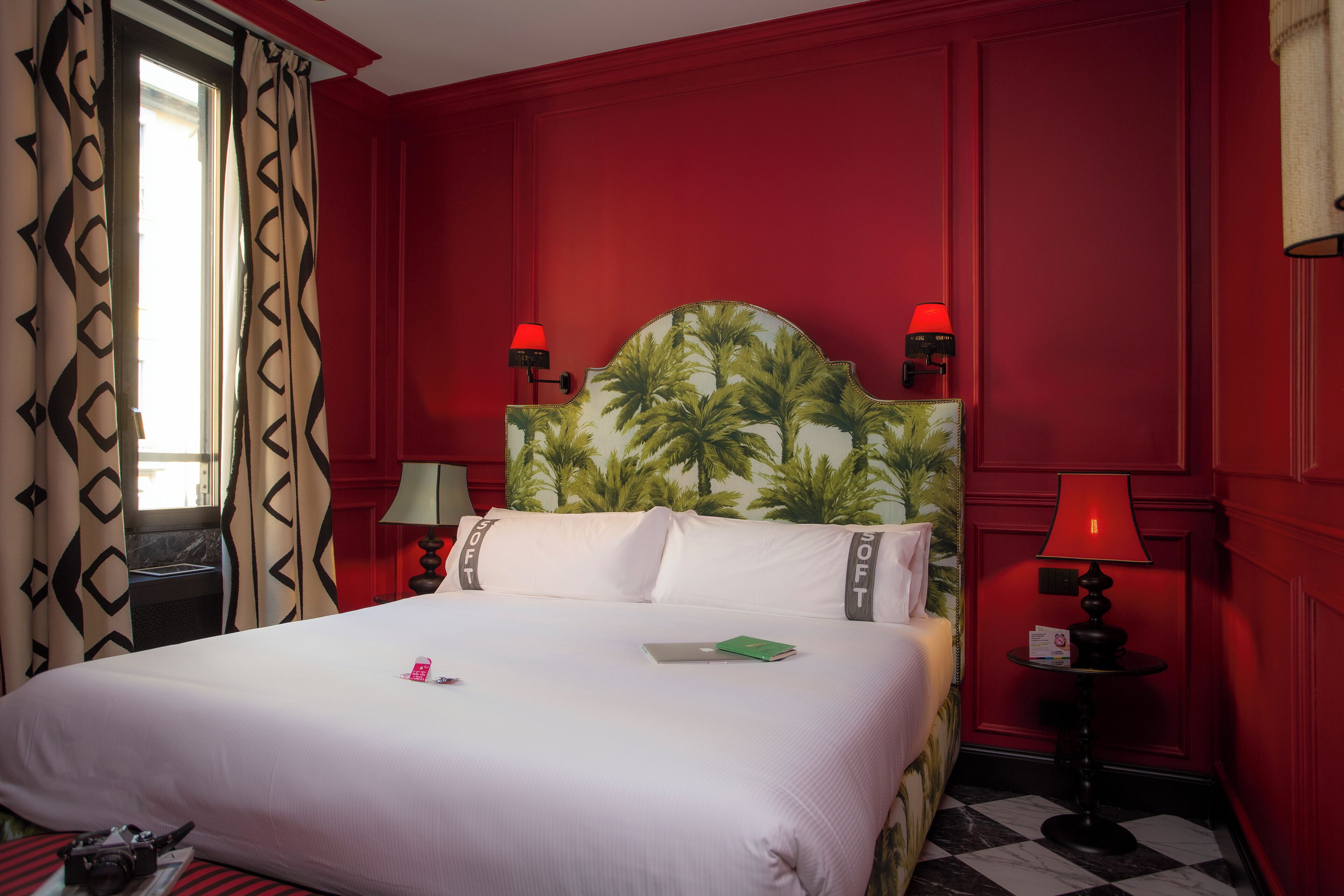 Habitación de hotel con pared roja, cama con ropa blanca, dos lámparas rojas y ventana con cortinas estampadas.