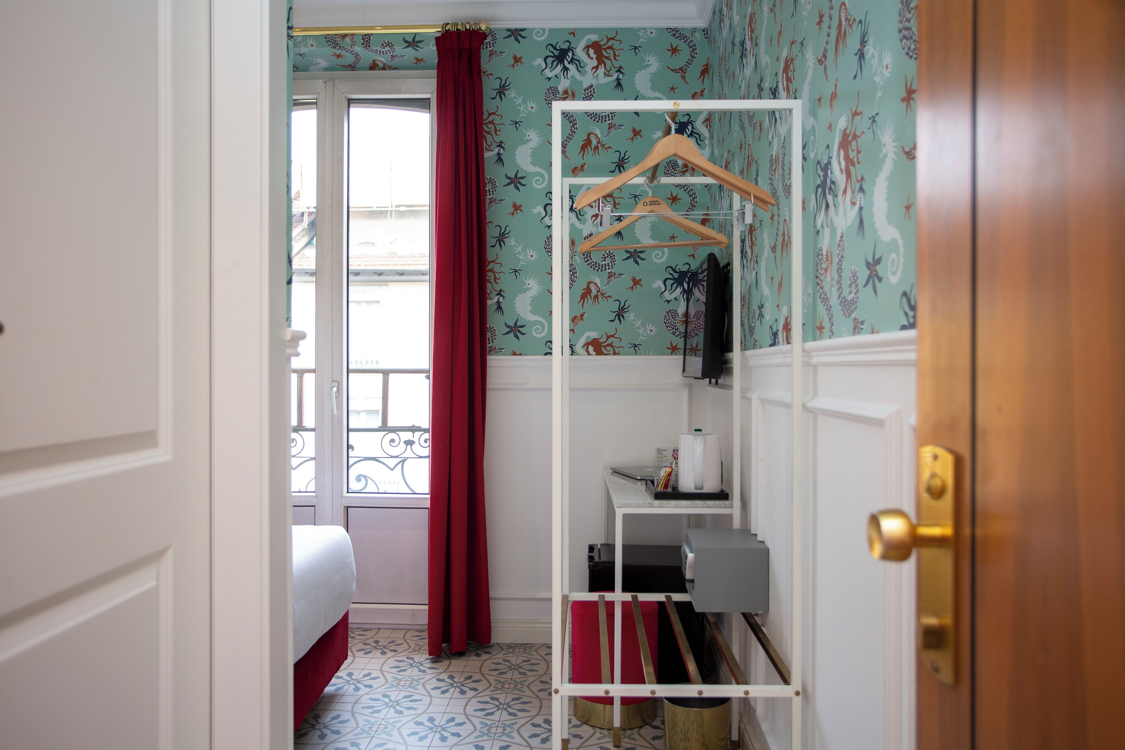 Una camera d'albergo con un letto, un armadio bianco, un appendiabiti e una finestra con tenda rossa.