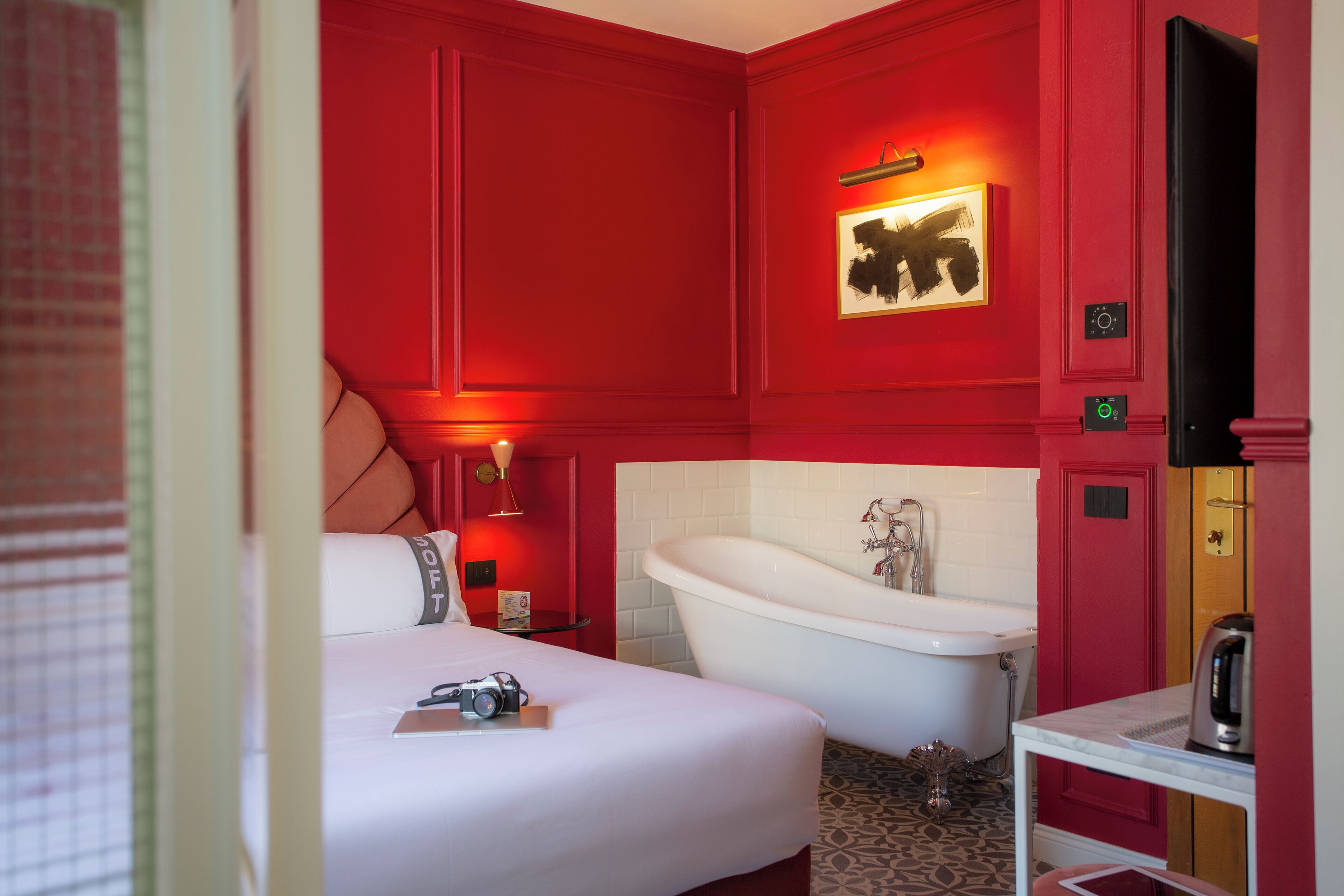 Una camera d'albergo con una parete rossa, una vasca da bagno bianca, un letto con una fotocamera sopra e un tavolino con un bollitore.