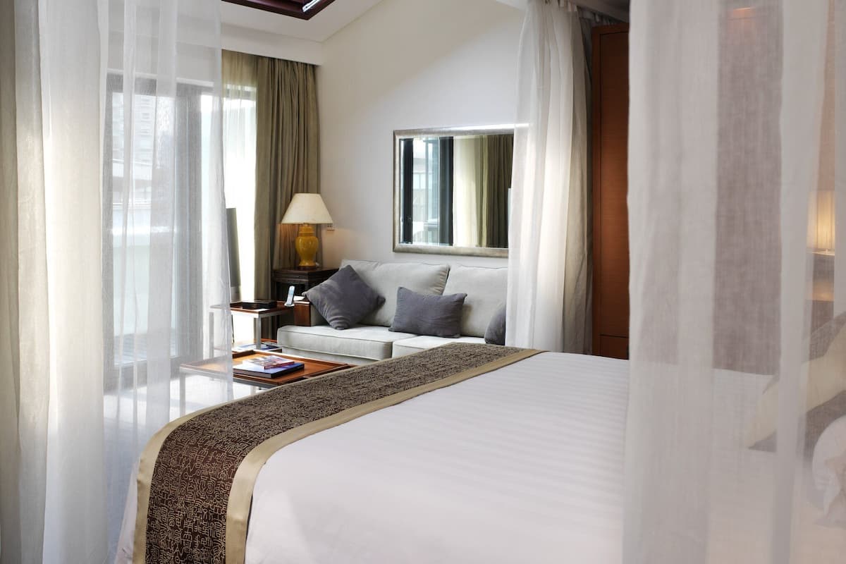 Una habitación de hotel con cama, mesita de noche, lámpara, espejo y cortinas translúcidas.