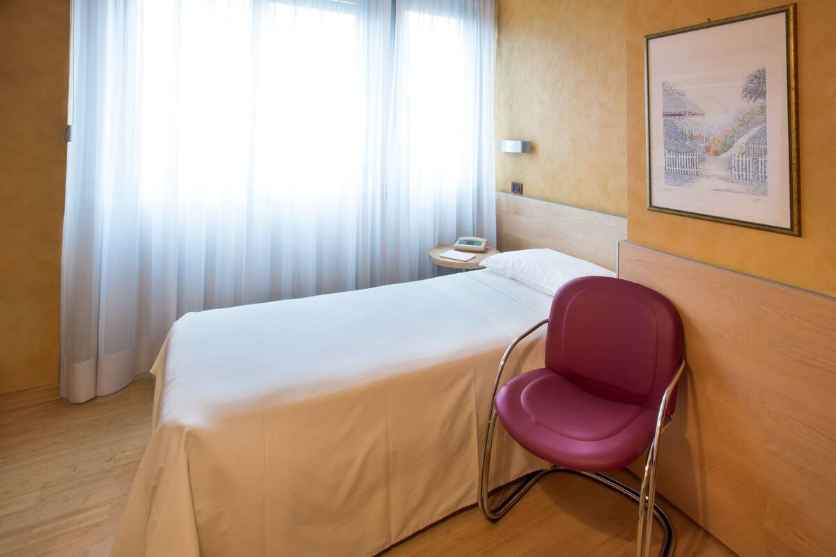 Una habitación de hotel con cama, una silla, una mesita de noche, una lámpara y un cuadro en la pared.