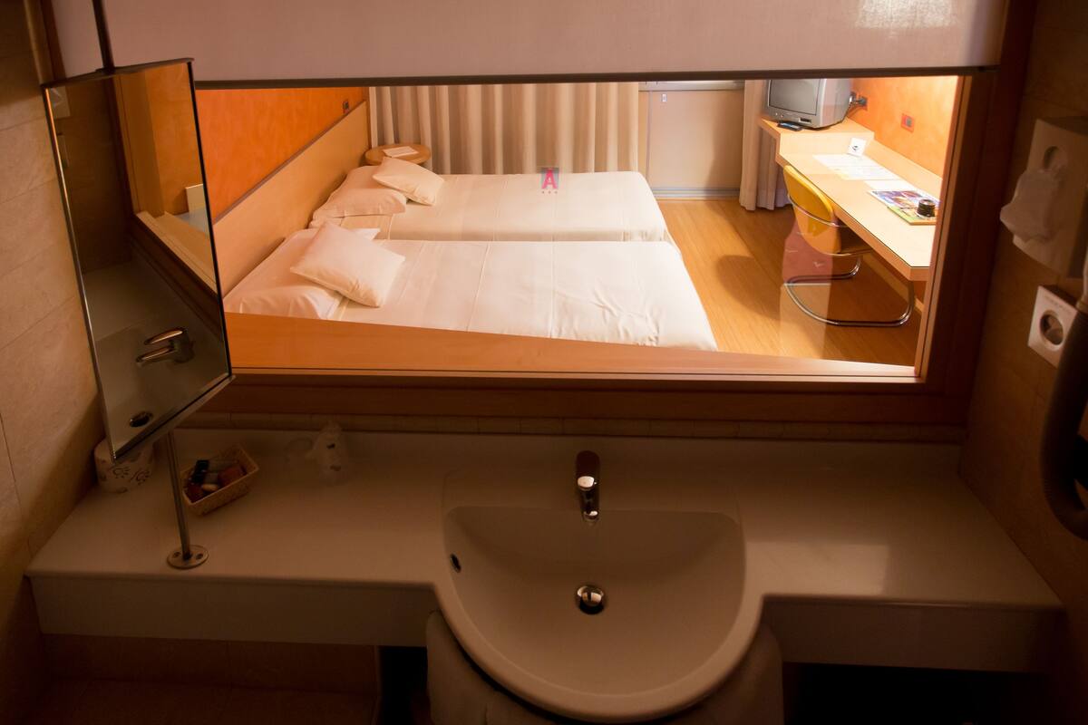 Una habitación de hotel con cama, mesita de noche, espejo, lavamanos y ventana.
